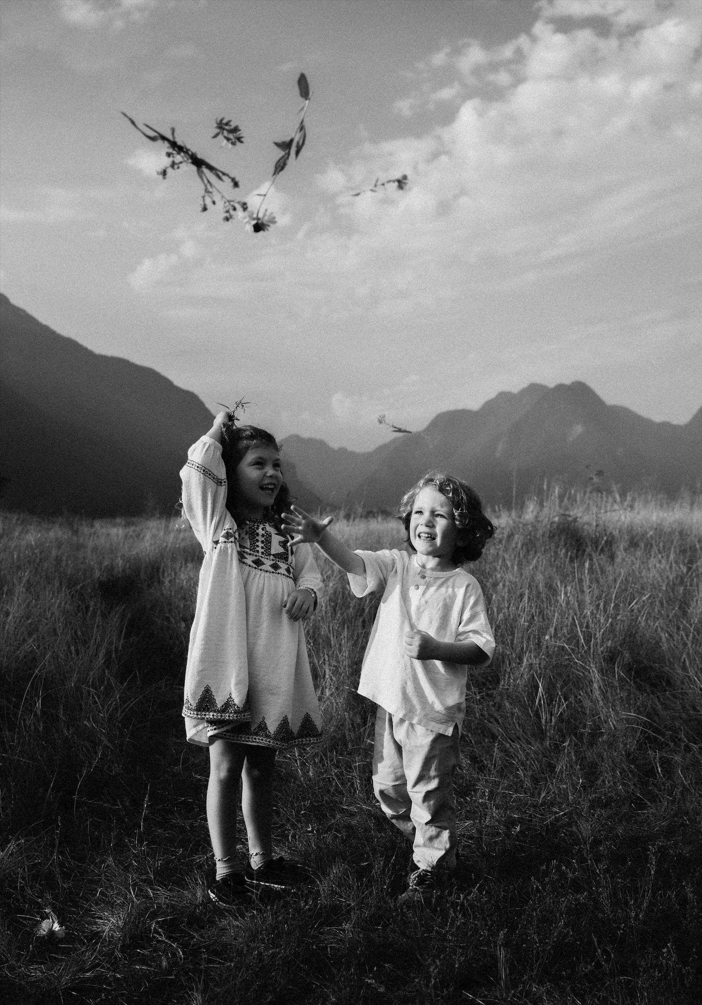 Two children, a girl and a boy, playing outdoors in a grassy field with mountains in the background. The girl is tossing a bunch of leaves or flowers into the air, and both are smiling and enjoying the moment.
