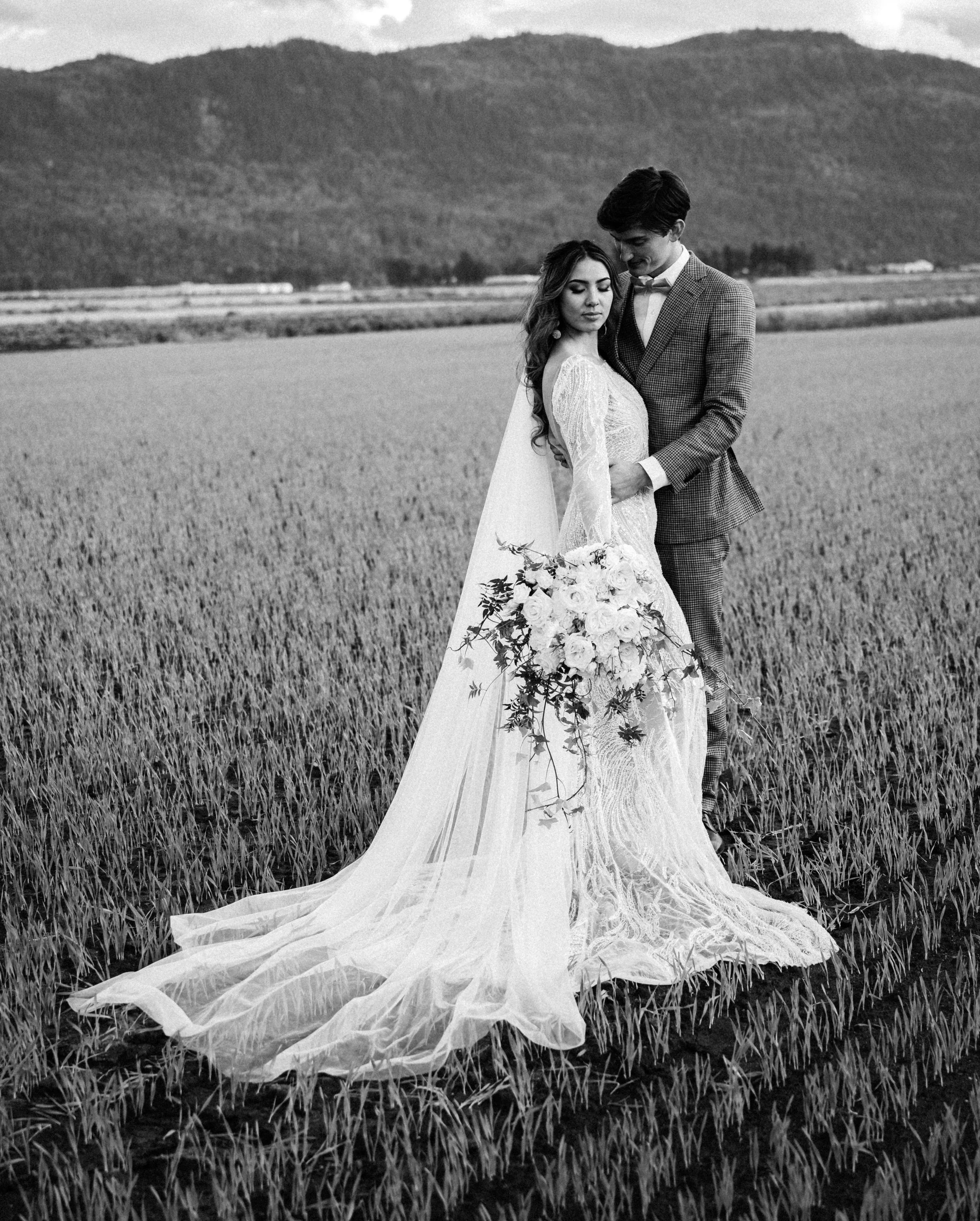 Black and white photo of a christian bride in a lace wedding gown with a long veil, holding a bouquet of flowers, standing with a groom in a suit in a field, with mountains in the background.
