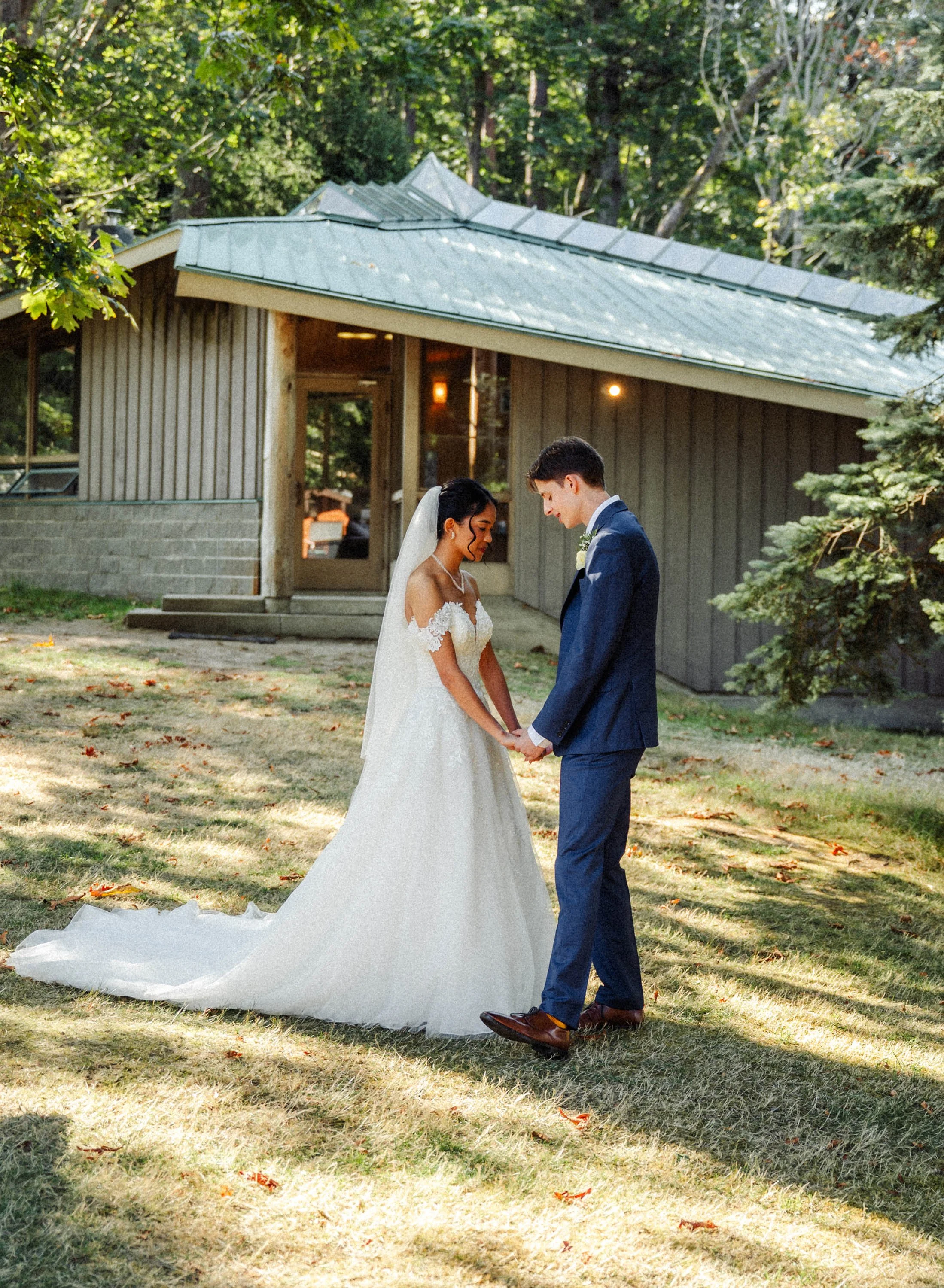 A bride and groom holding hands outside in a wooded area, with a small wooden building in the background, sharing a moment of prayer before the ceremony.