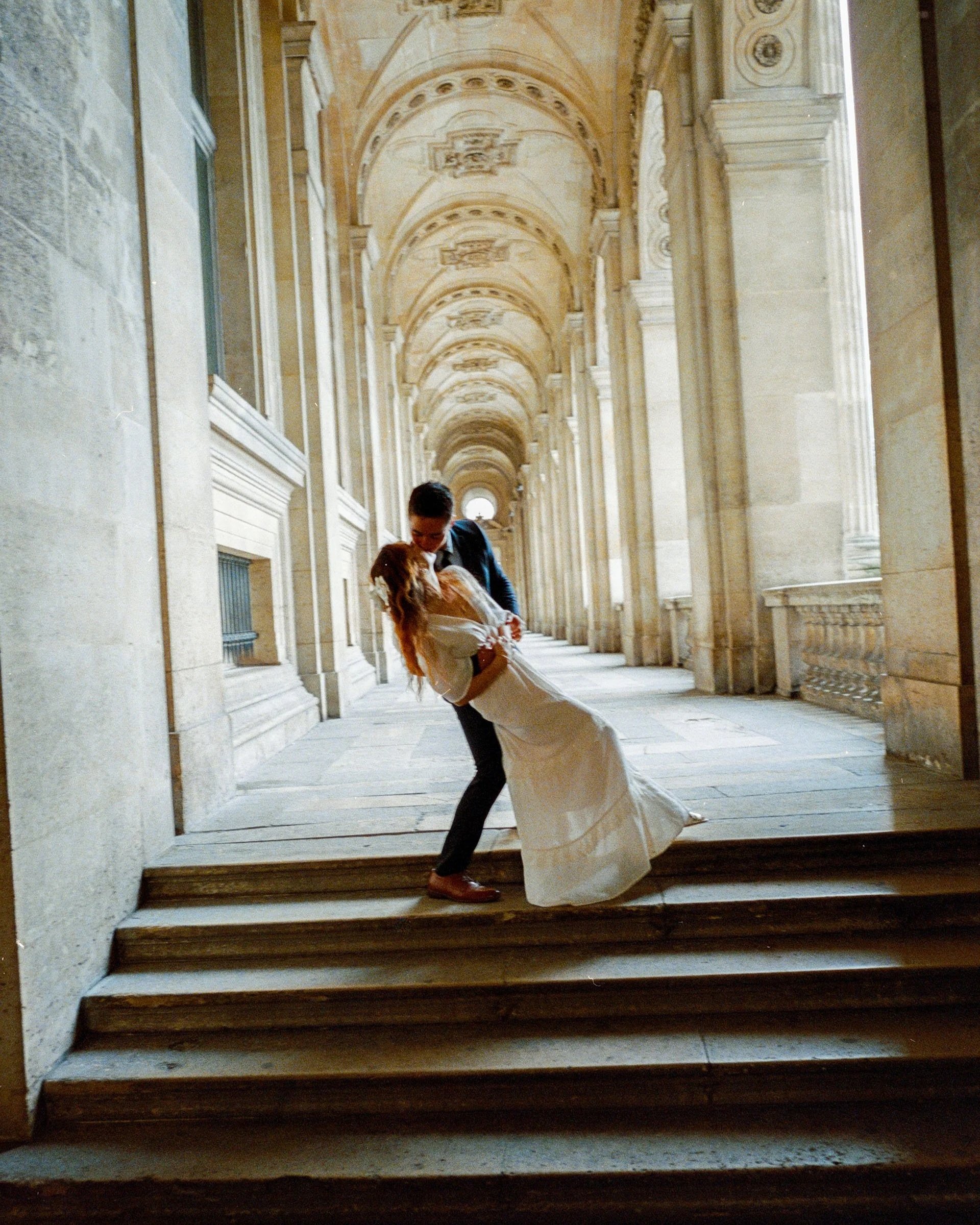 A couple dressed in wedding attire dancing on steps in an ornate, arched corridor of a historic building.