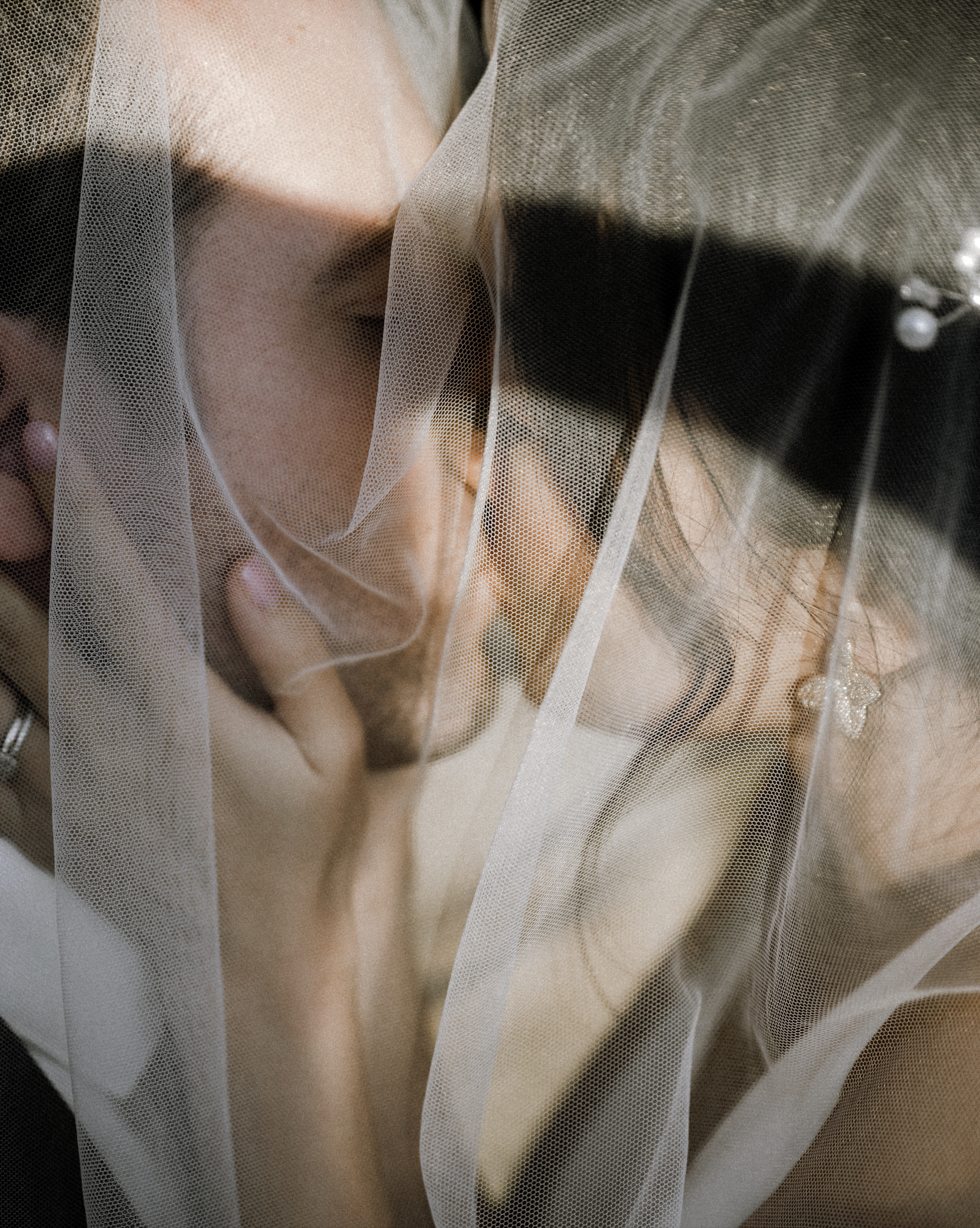 Two women are close together behind a sheer fabric veil, with one woman kissing the other's forehead and both women having their eyes closed.
