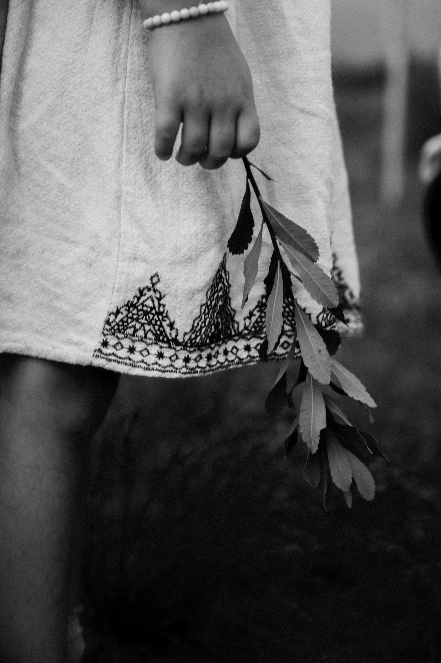 Black and white photo of a child holding a small branch with leaves, wearing a dress with detailed embroidery and a bracelet.