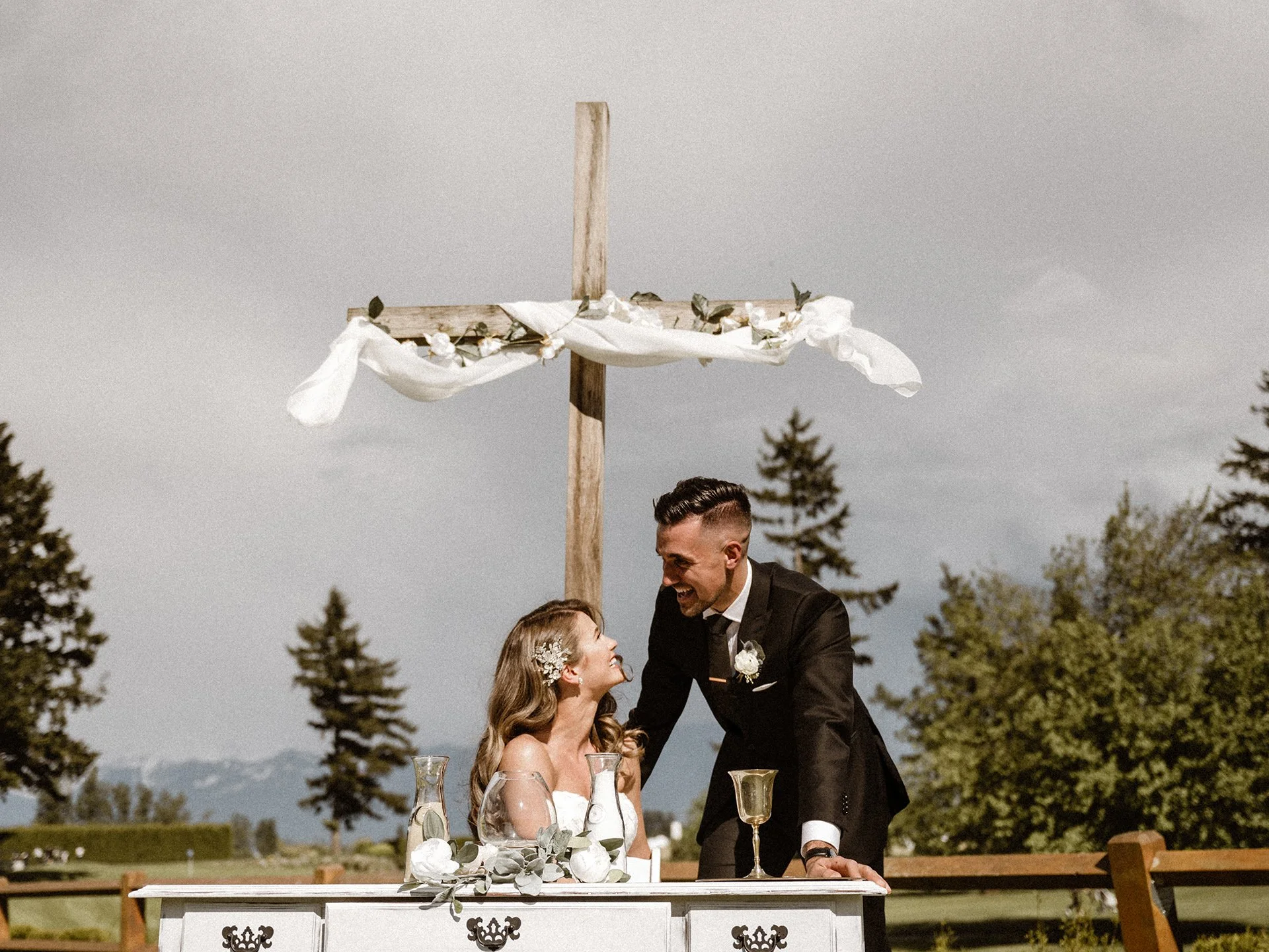 A wedding reception outdoors with a bride and groom smiling at each other, sitting at a white table with glasses and flower decorations. The groom, dressed in a black tuxedo, is leaning over the bride, who has long hair with floral accessories. In the background, there are trees and mountainous scenery under a cloudy sky, with a wooden cross decorated with white fabric and greenery behind them.