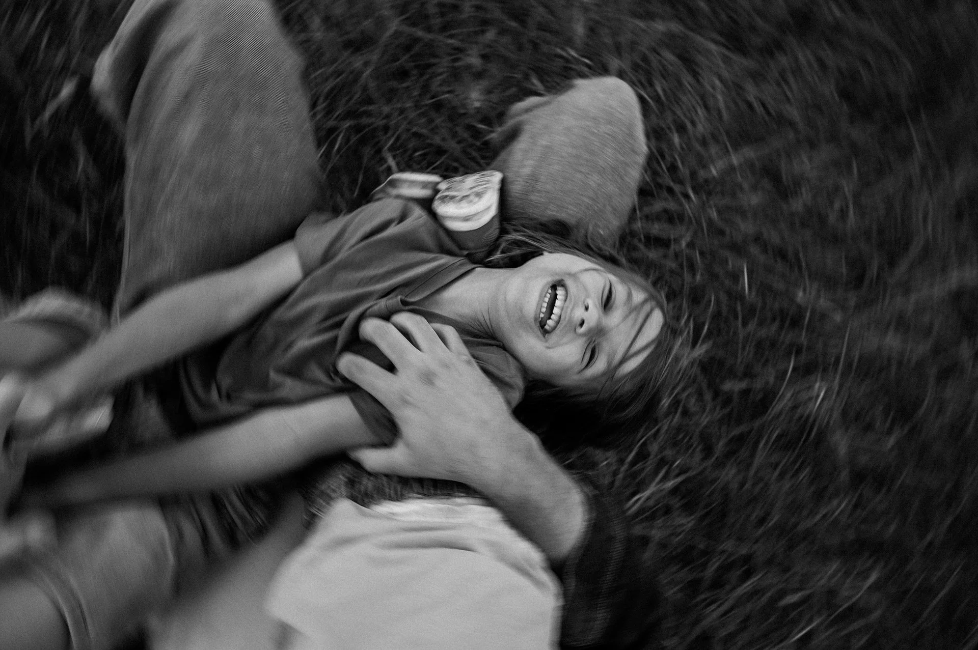 A child with long hair smiling and lying on grass, being played with by an adult whose hand is on the child's chest.