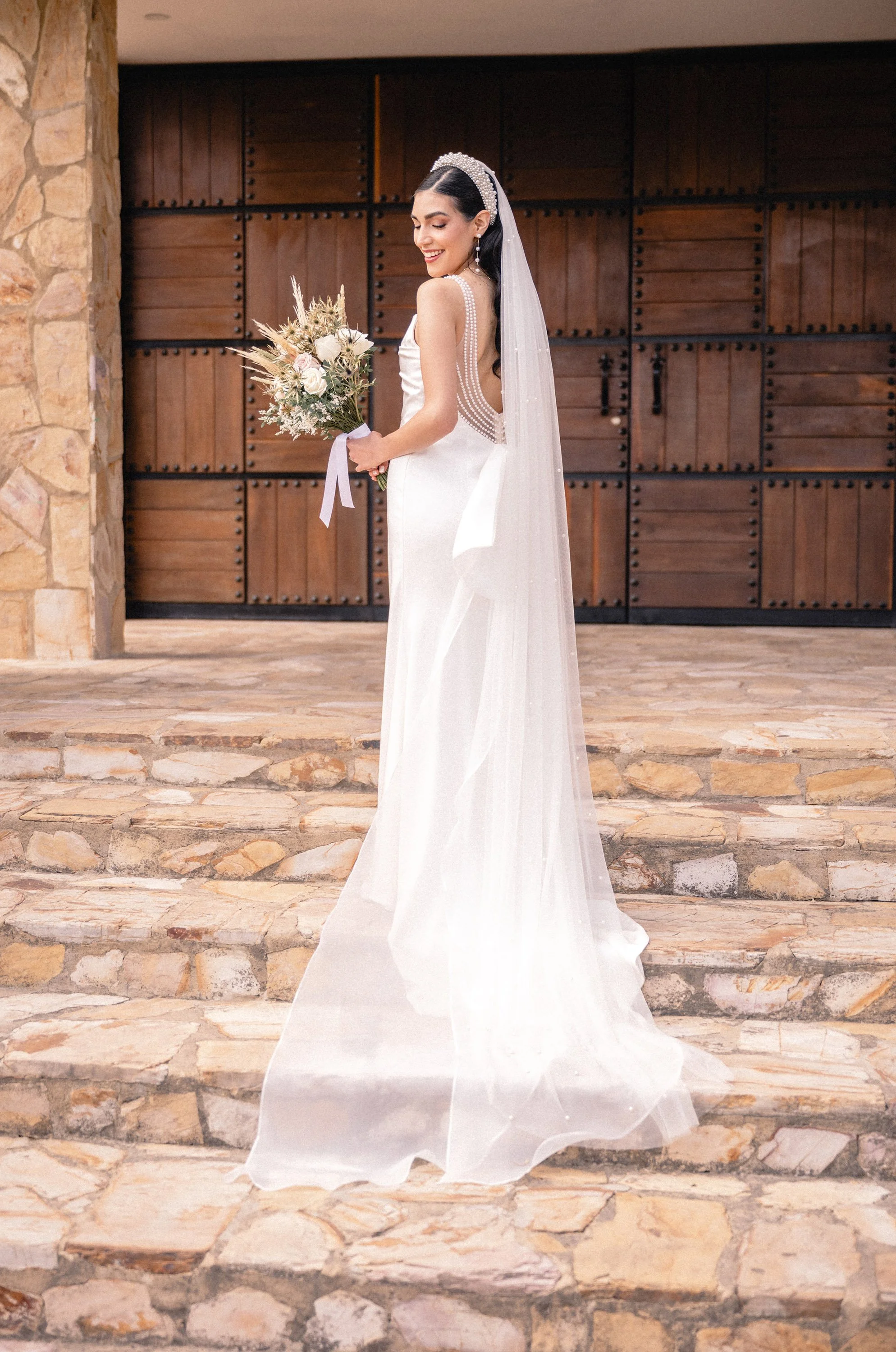 A christian bride in a white wedding dress holding a bouquet of flowers, standing on a stone staircase outside a building with large wooden doors.