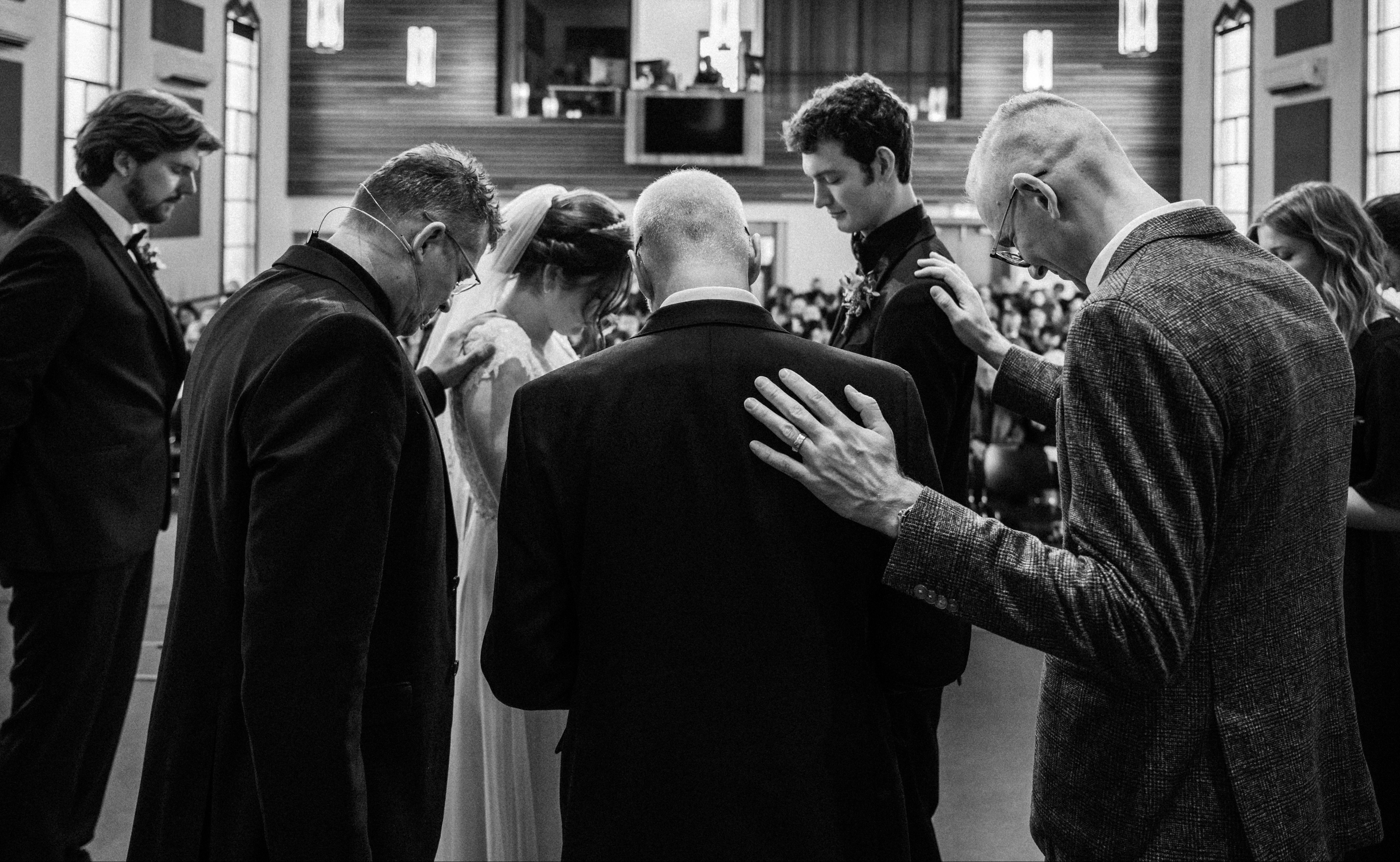 A black and white photo of a group of people praying or reflecting with heads bowed and hands on each other's shoulders inside a church during a wedding ceremony.