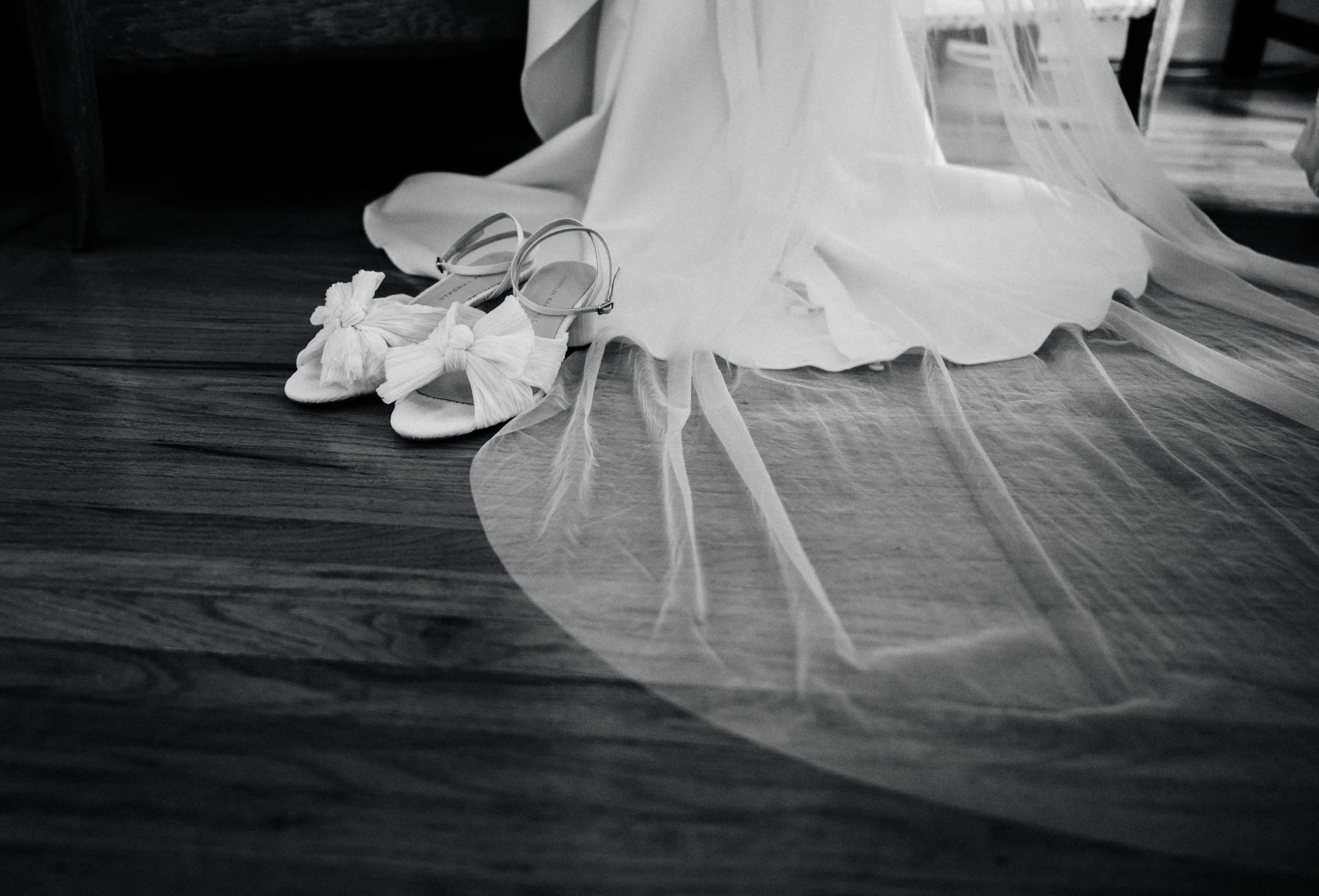 Black and white photograph of a pair of embellished, open-toe heels with bows, a wedding dress with tulle fabric, and a piece of furniture with a napkin or cloth on top, all placed on a wooden floor.