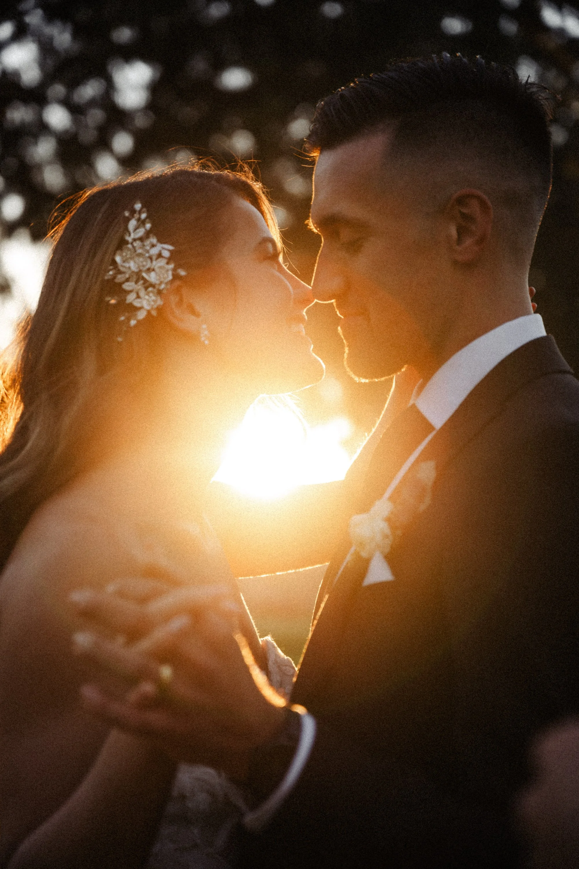 A couple at their wedding, embracing each other and smiling with their noses touching, backlit by sunset, with blurred trees in the background.