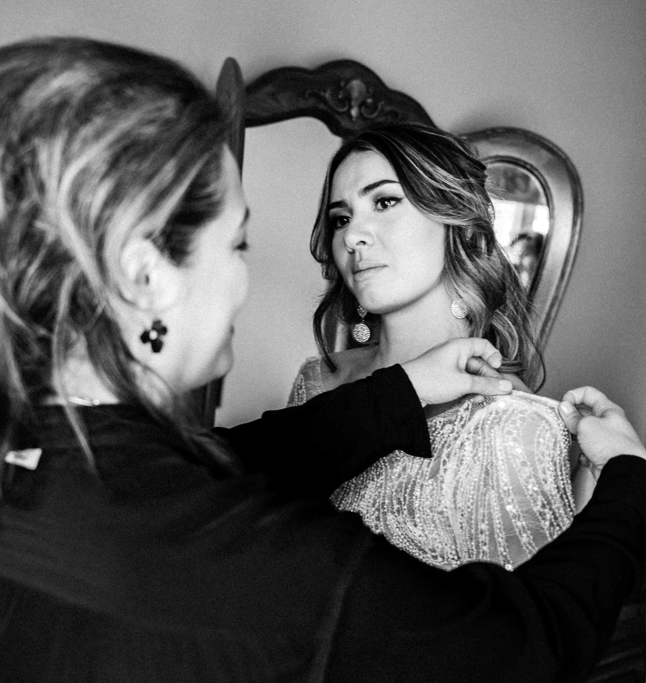 bride's mother is adjusting the shoulder of the bride, who is looking at her with a serious expression. The scene takes place in a room with a mirror in the background.