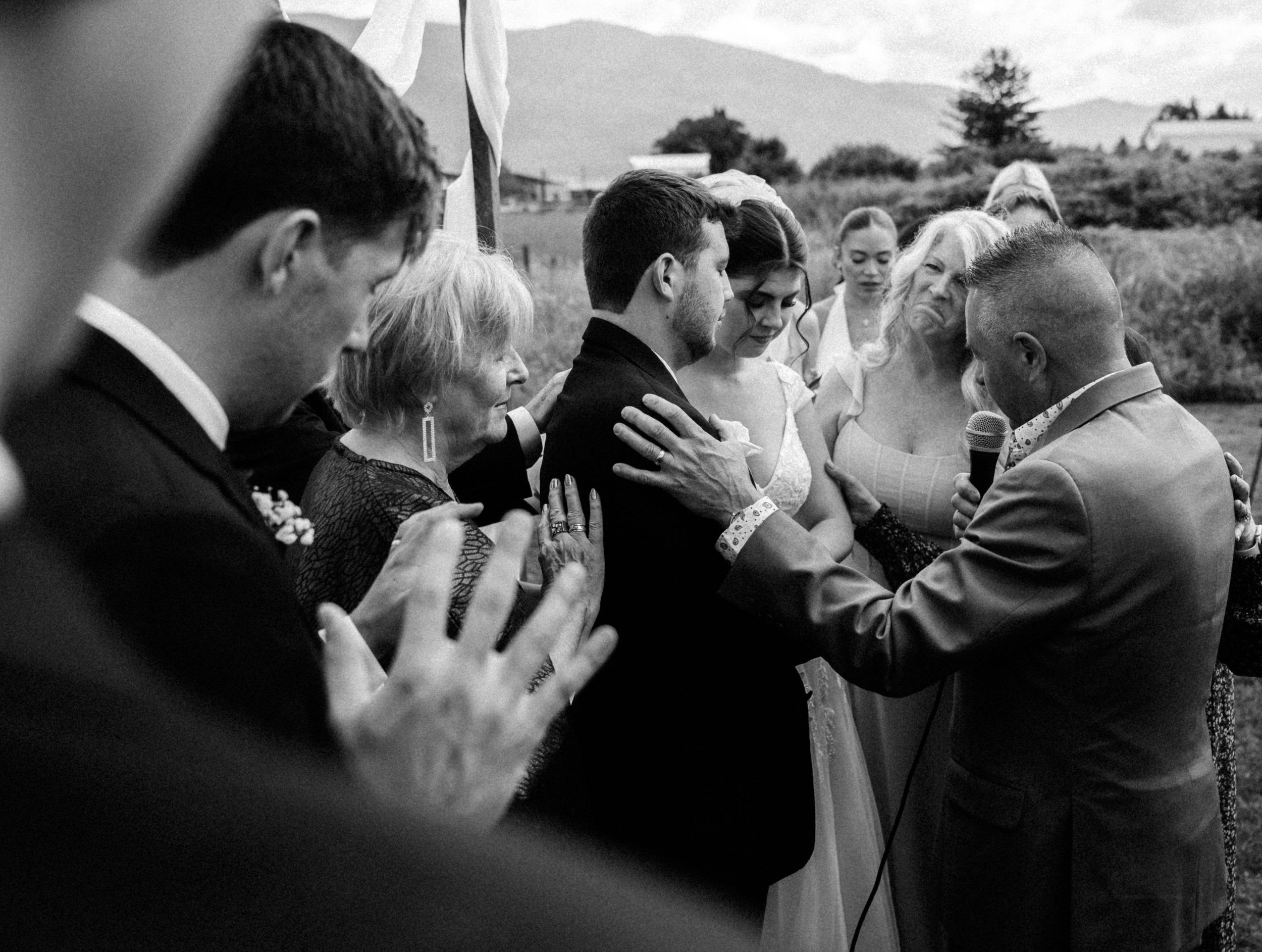 Black and white photo of a wedding ceremony outdoors with the couple and family with their heads bowed and hands on each other, and a man with a microphone officiating.