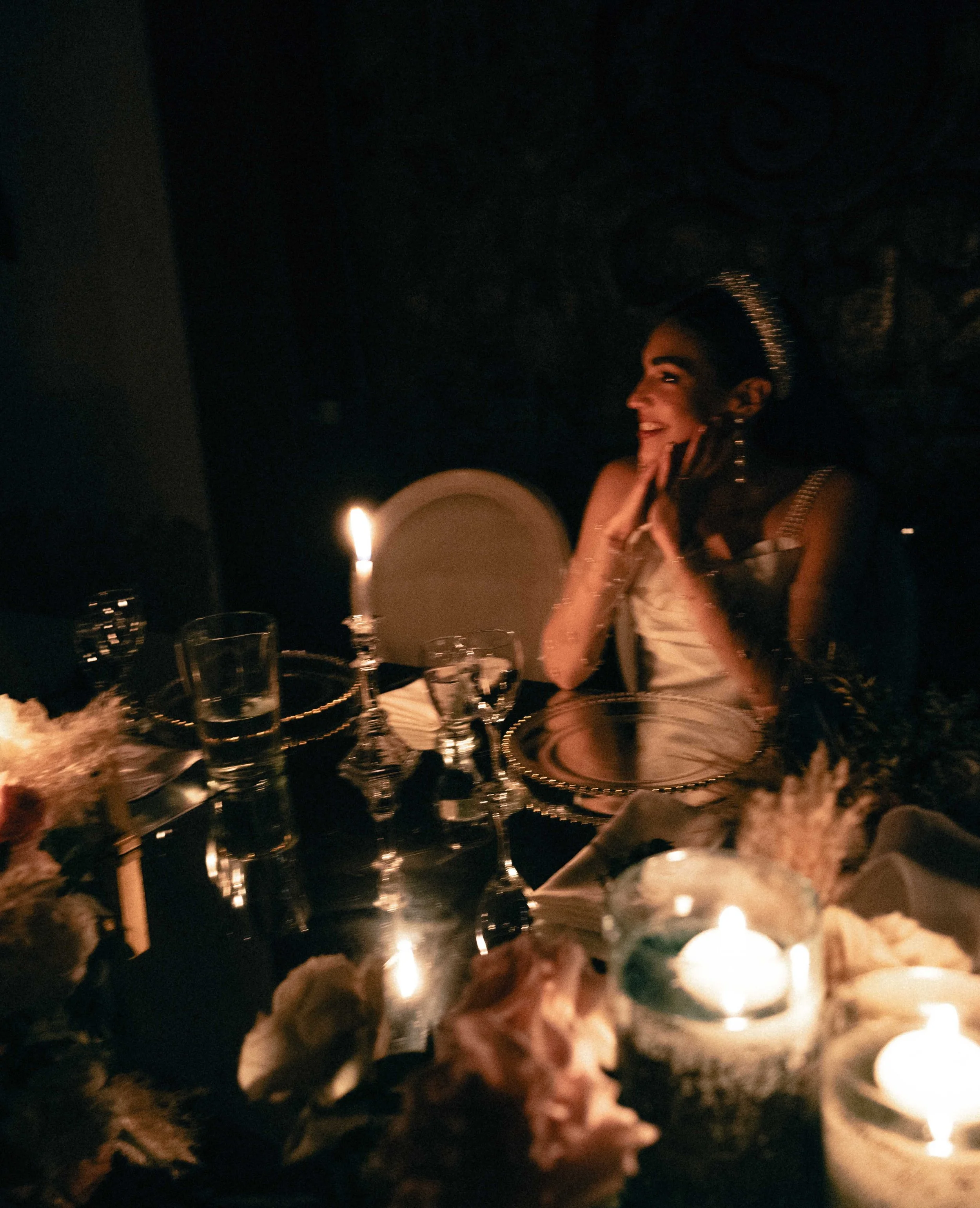 A christian bride with a crown, smiling, sitting at a dimly lit table with candles and glasses, in an elegant setting.