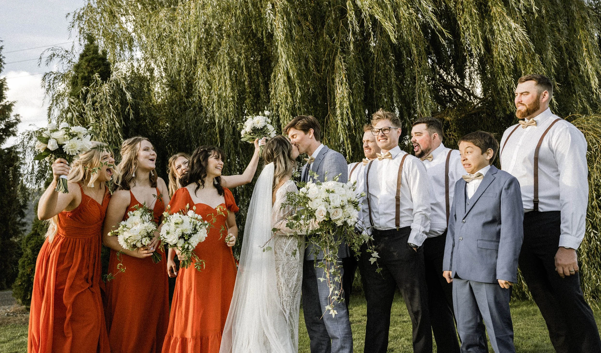 A wedding outdoor ceremony with a bride and groom kissing under a large tree surrounded by bridesmaids in orange dresses holding bouquets and groomsmen in white shirts, suspenders, and bow ties, all smiling and celebrating.