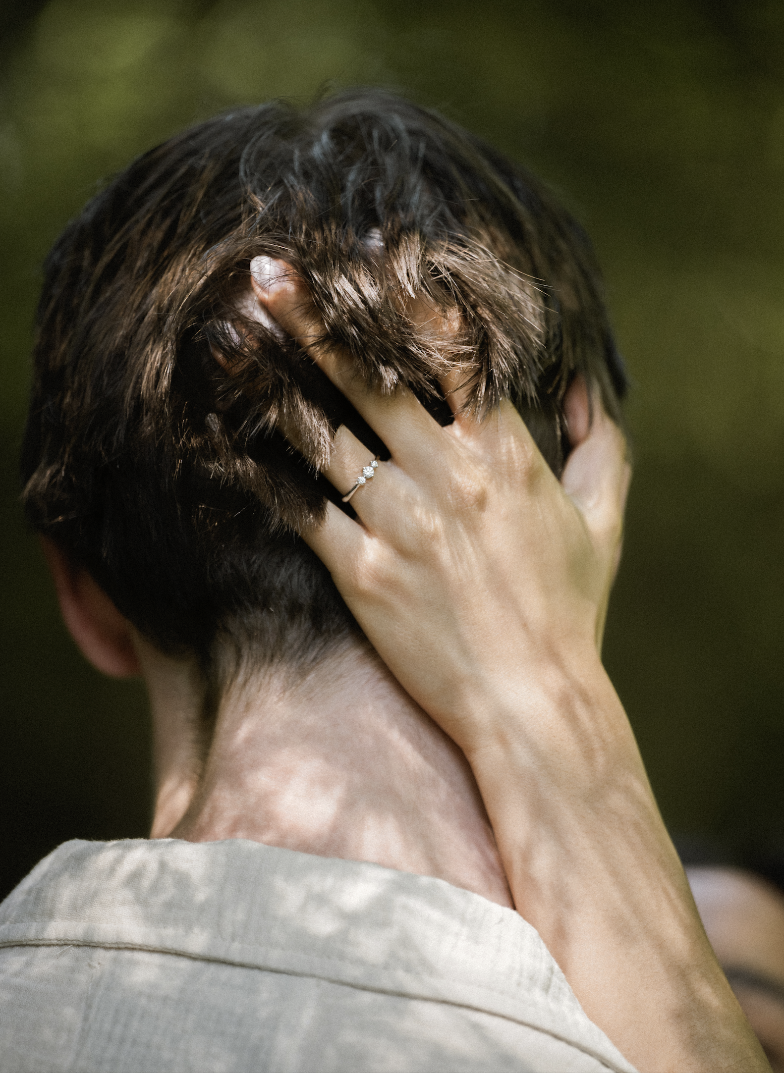 A woman with short brown hair is holding her head with her hands, covering her face. She is wearing a ring on her finger and a light-colored top. The background is blurred with green tones.