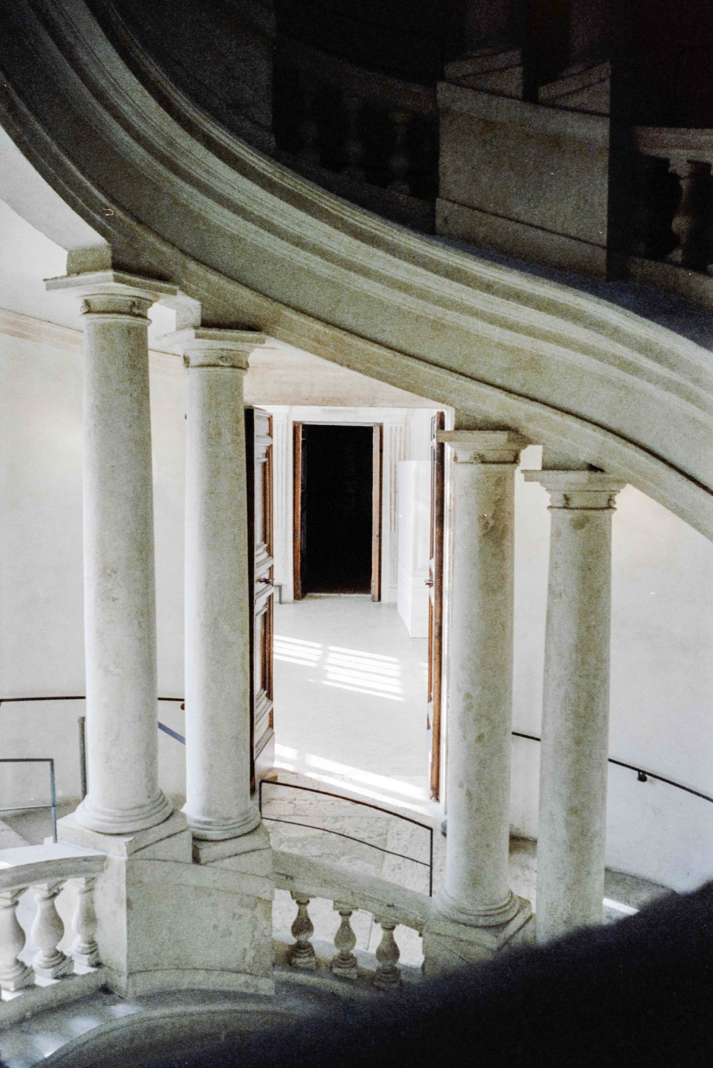 Interior view of a grand staircase with ornate columns, leading to a doorway with sunlight streaming through open doors.