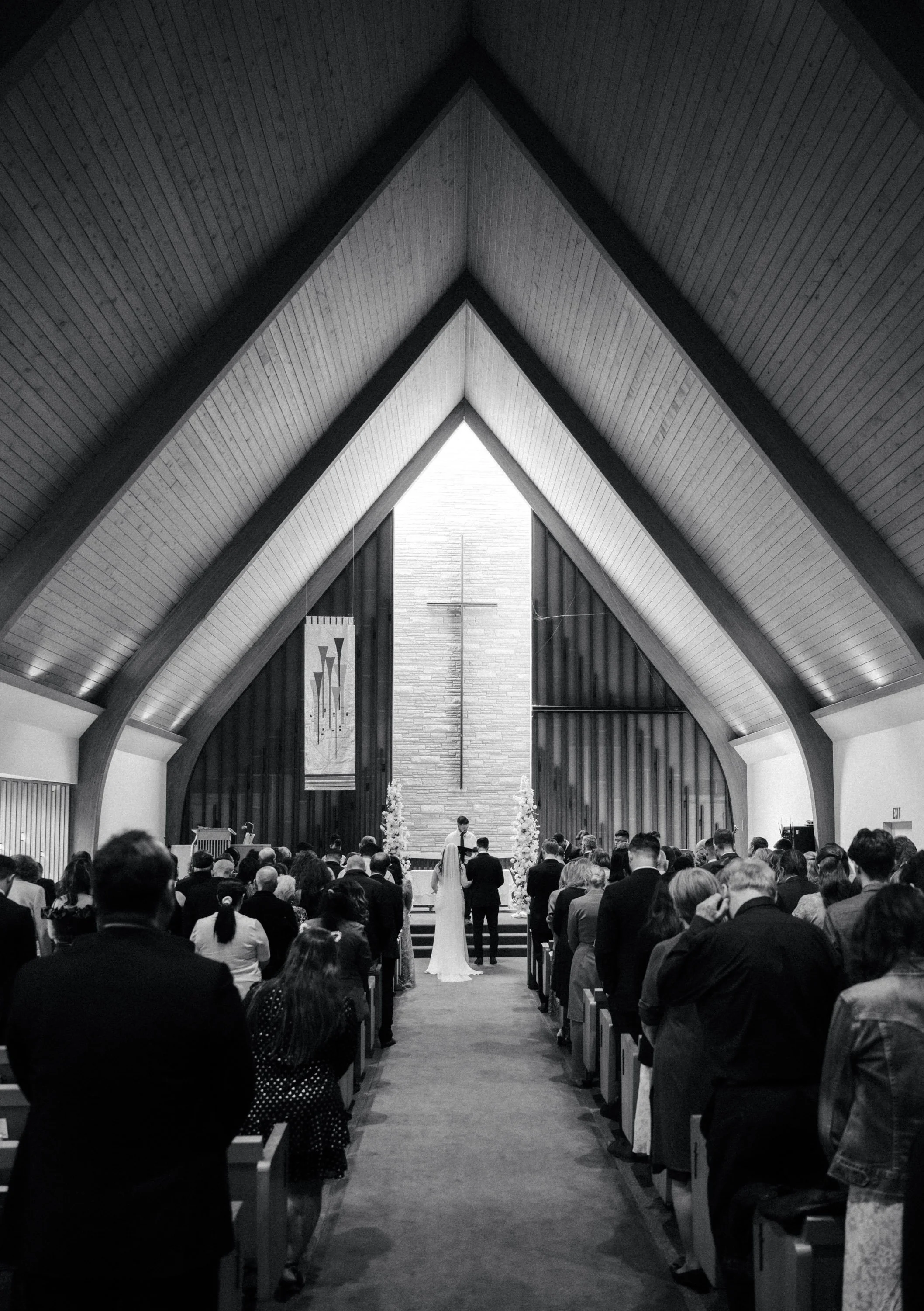 Interior of a church with a high, arched, wooden ceiling, congregation members seated, a couple standing at the altar during a wedding ceremony, a cross on the wall behind them, and banners hanging on either side.
