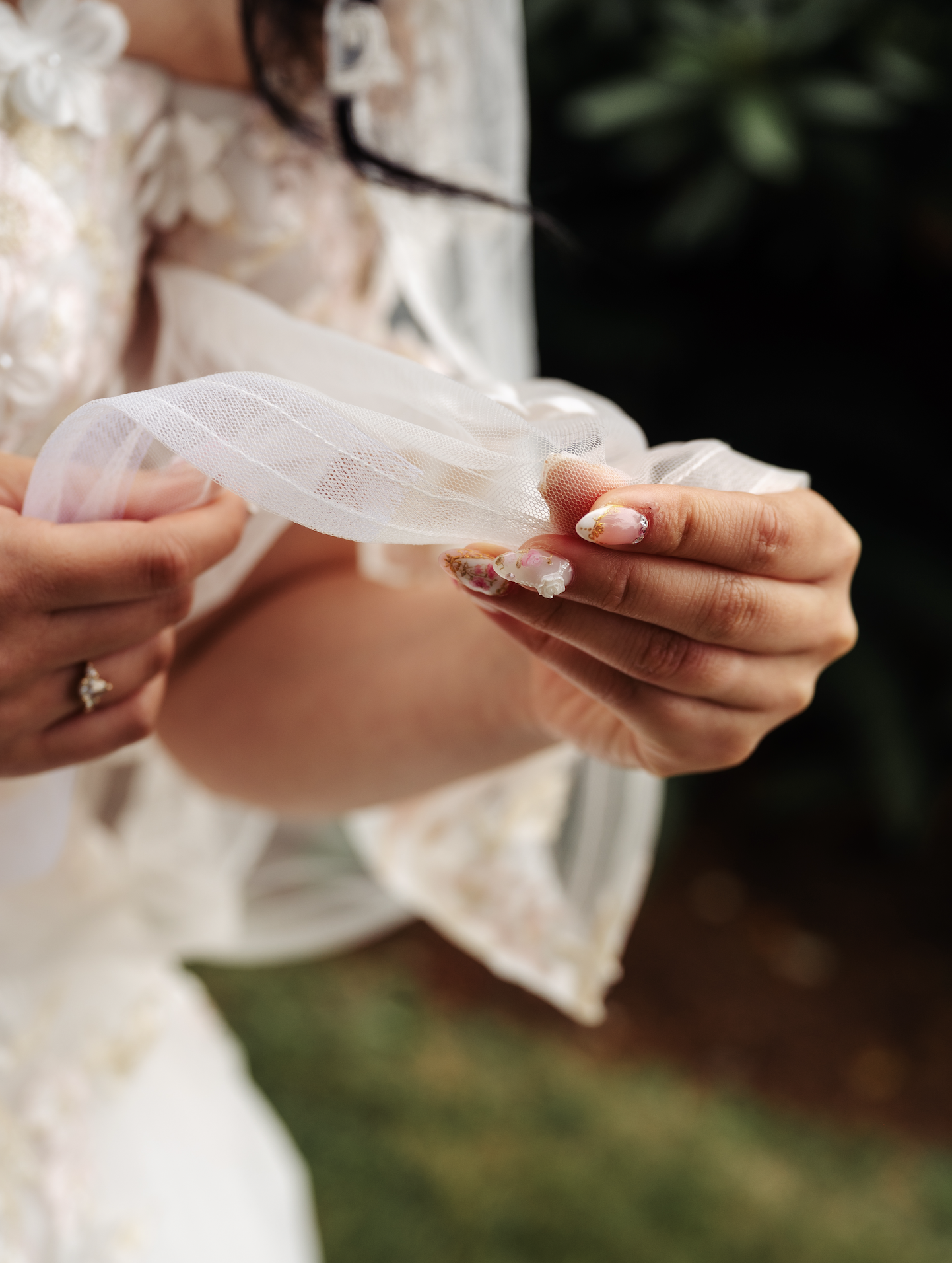 A person holding a piece of white tulle fabric, with detailed floral nail art and a wedding ring, outdoors with blurred greenery in the background.
