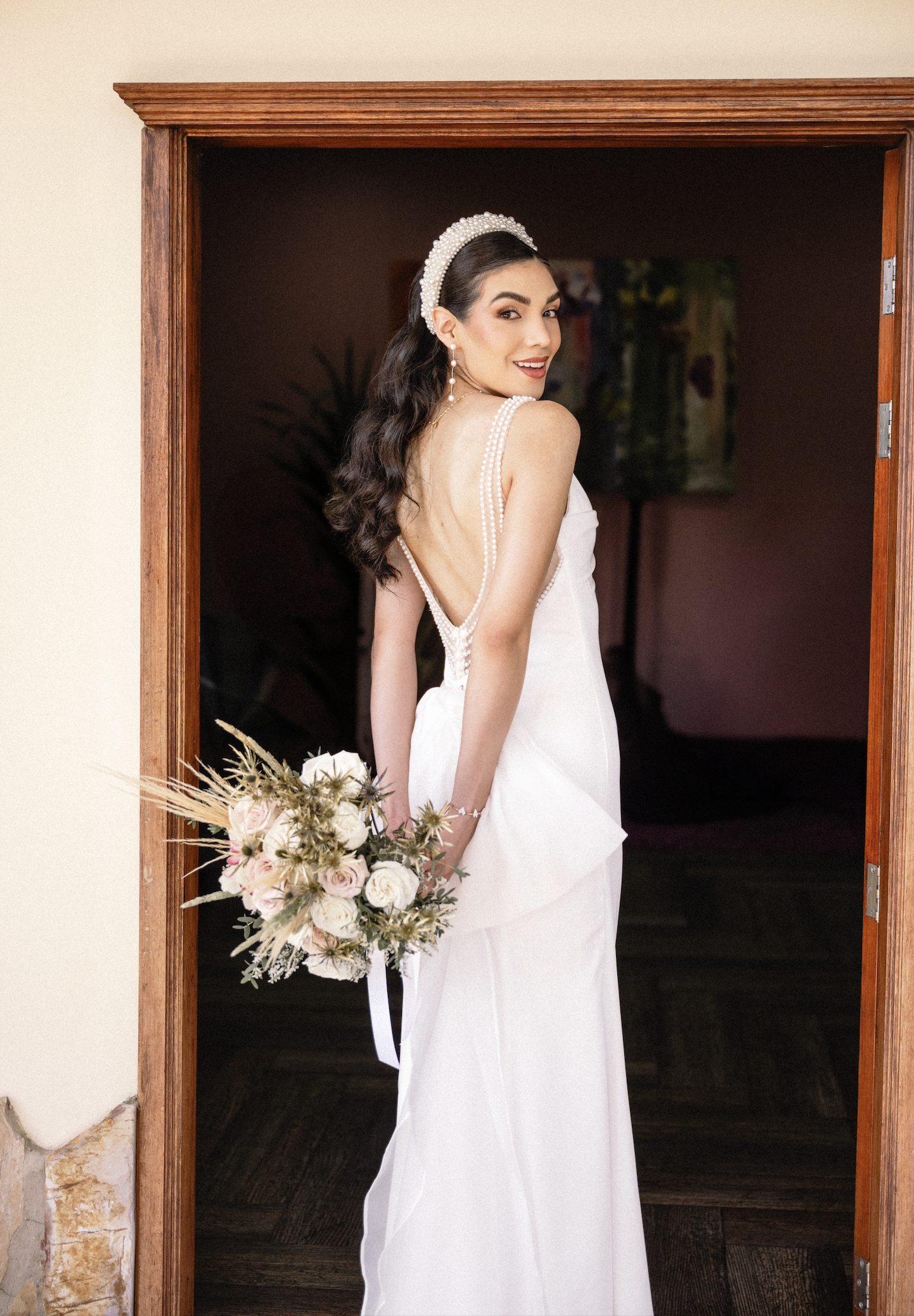 A woman in a white wedding dress with pearl accents, holding a bouquet of pink and white flowers, standing in a doorway, smiling and looking over her shoulder.