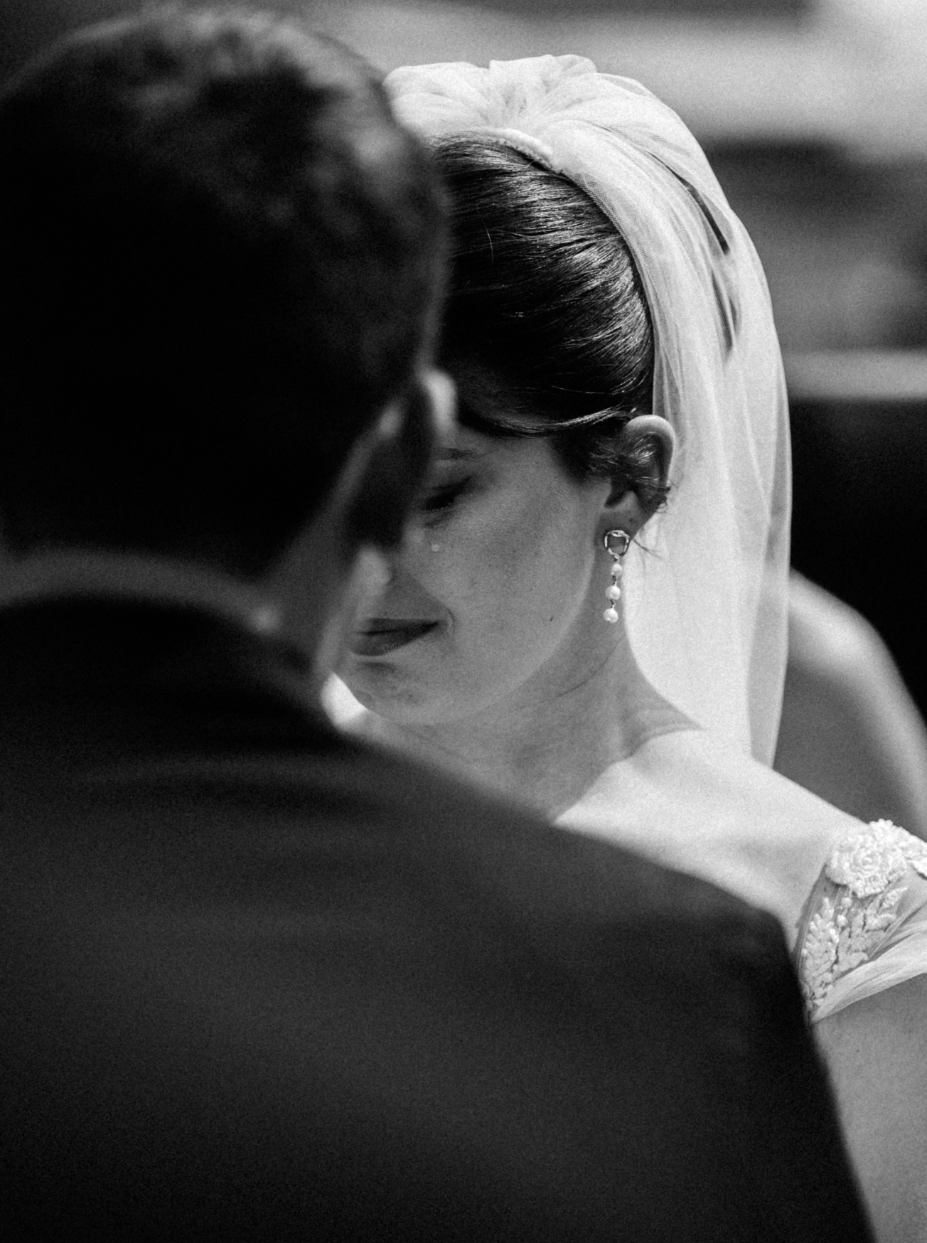 A black and white photo of a bride and groom in a close embrace, with the bride wearing earrings and a veil, and the groom's back partially visible.