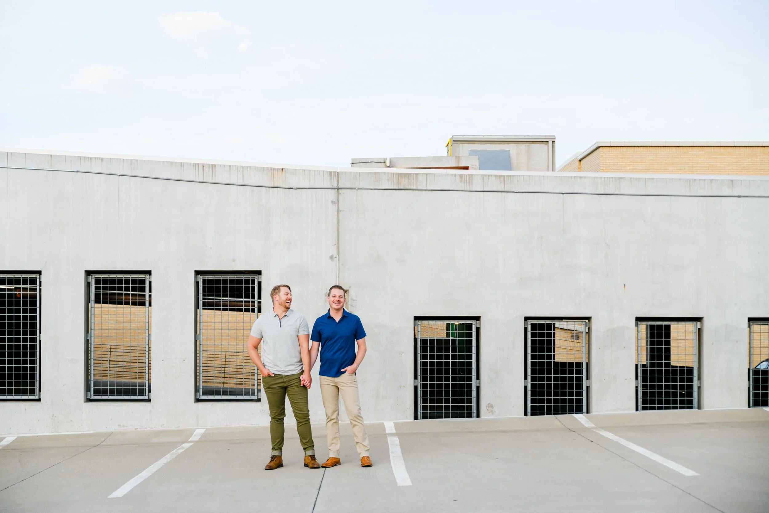 Two men standing and holding hands in a parking lot, smiling and talking, against a concrete building with barred windows for their gay LGBTQ+ denver proposal photos