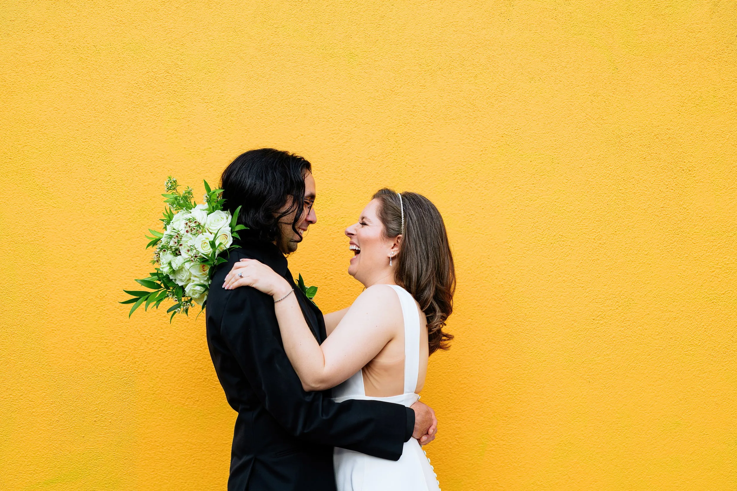 Couple laughs together on their art gallery wedding day against a yellow wall in downtown Denver, captured by Catherine Norwood Photography