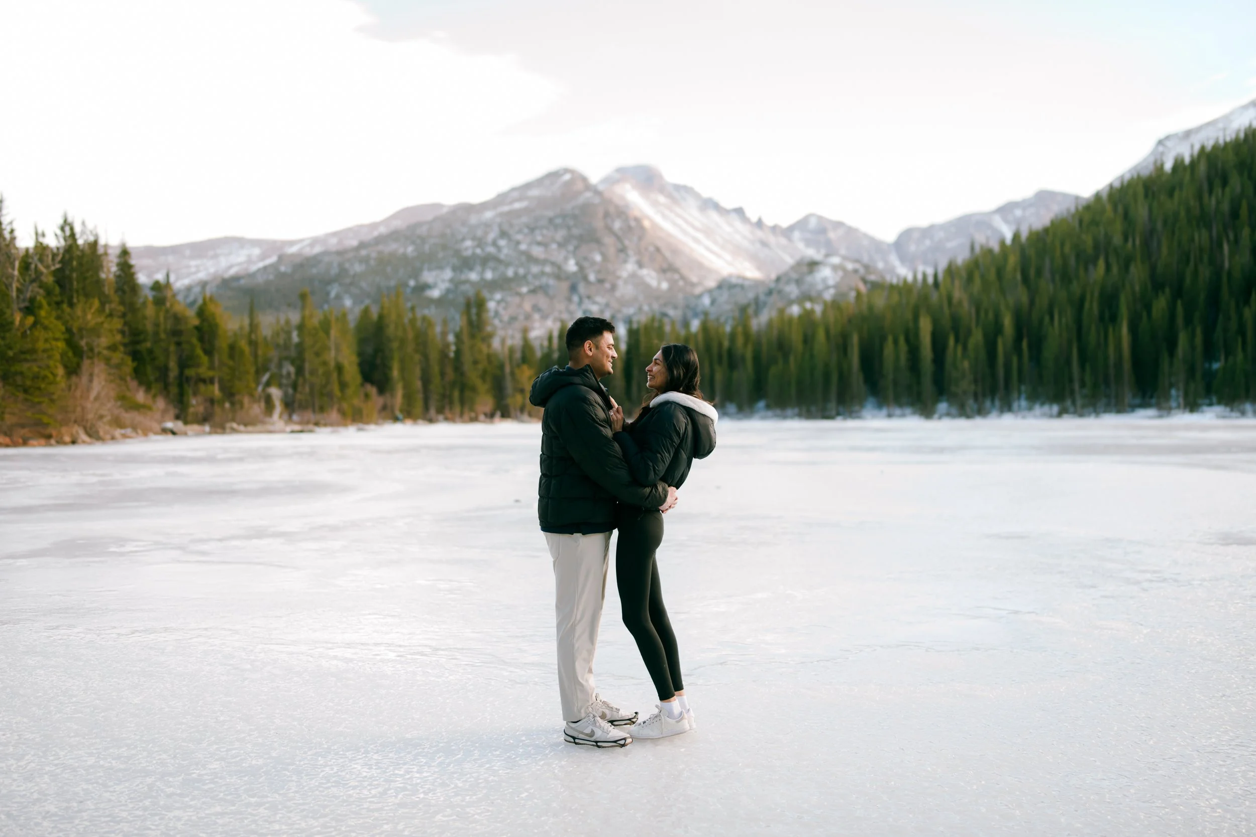 A BIPOC Indian couple stands on a frozen Bear Lake in the Winter for their Rocky Mountain National Park Colorado proposal photography. They're facing each other and smiling, with mountains and evergreen trees in the background.