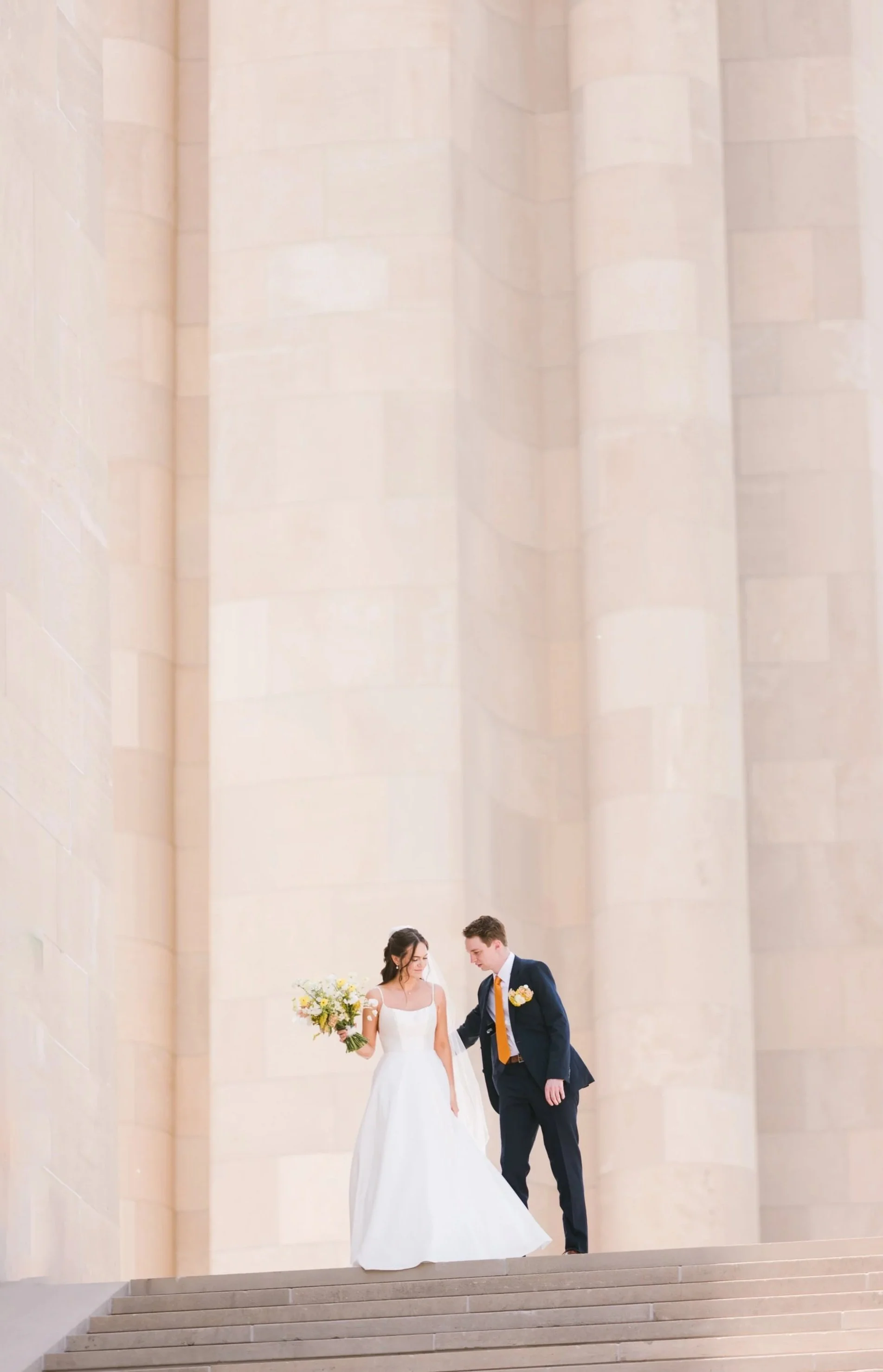 A bride and groom walk down steps in front of large stone columns, the bride carrying a bouquet — a timeless scene for modern kansas city wedding photography.