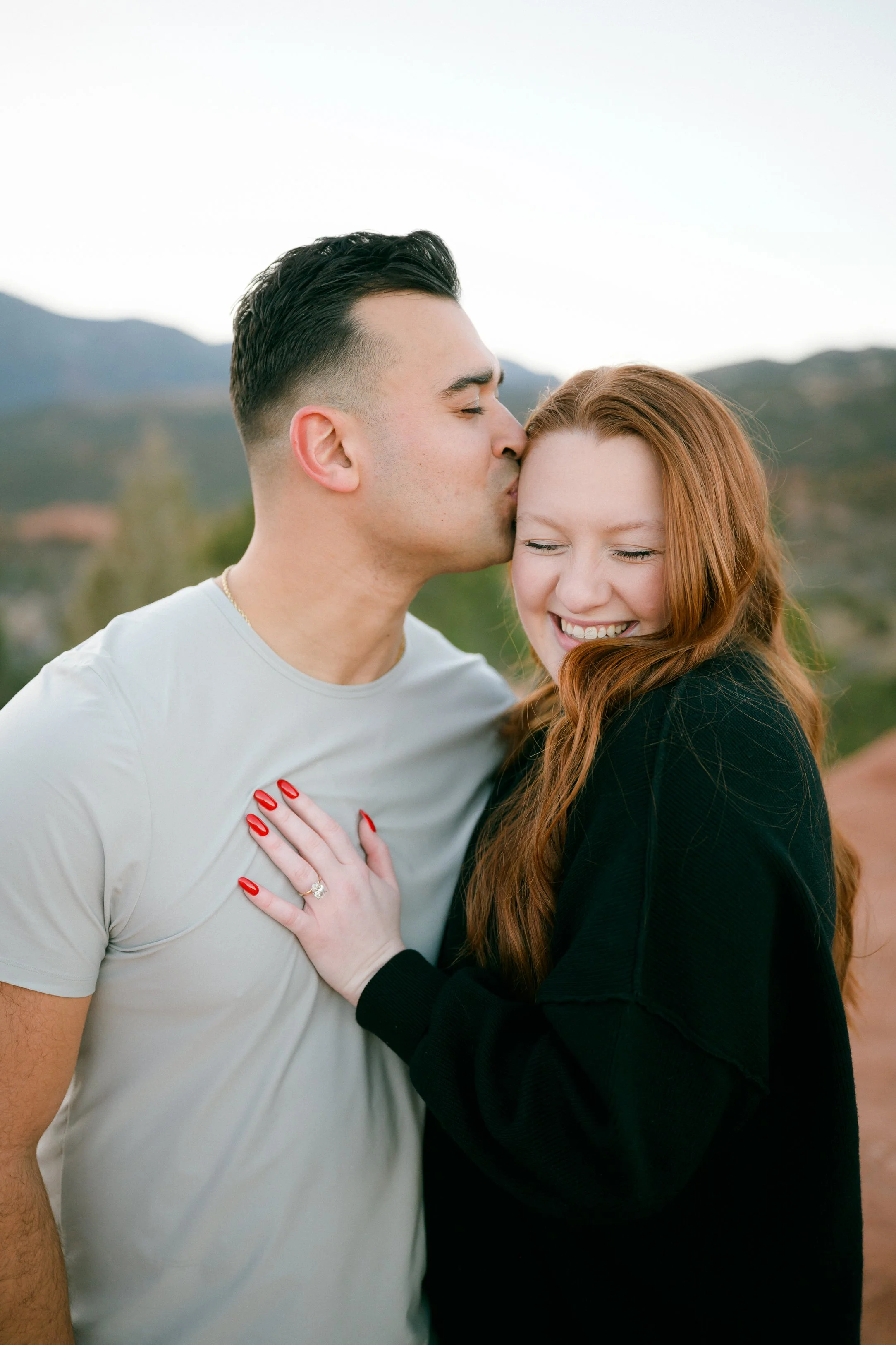 A man kissing a woman on the forehead while she smiles with her eyes closed and hand on his chest, outdoors with mountains in the background for their BIPOC colorado springs proposal photos