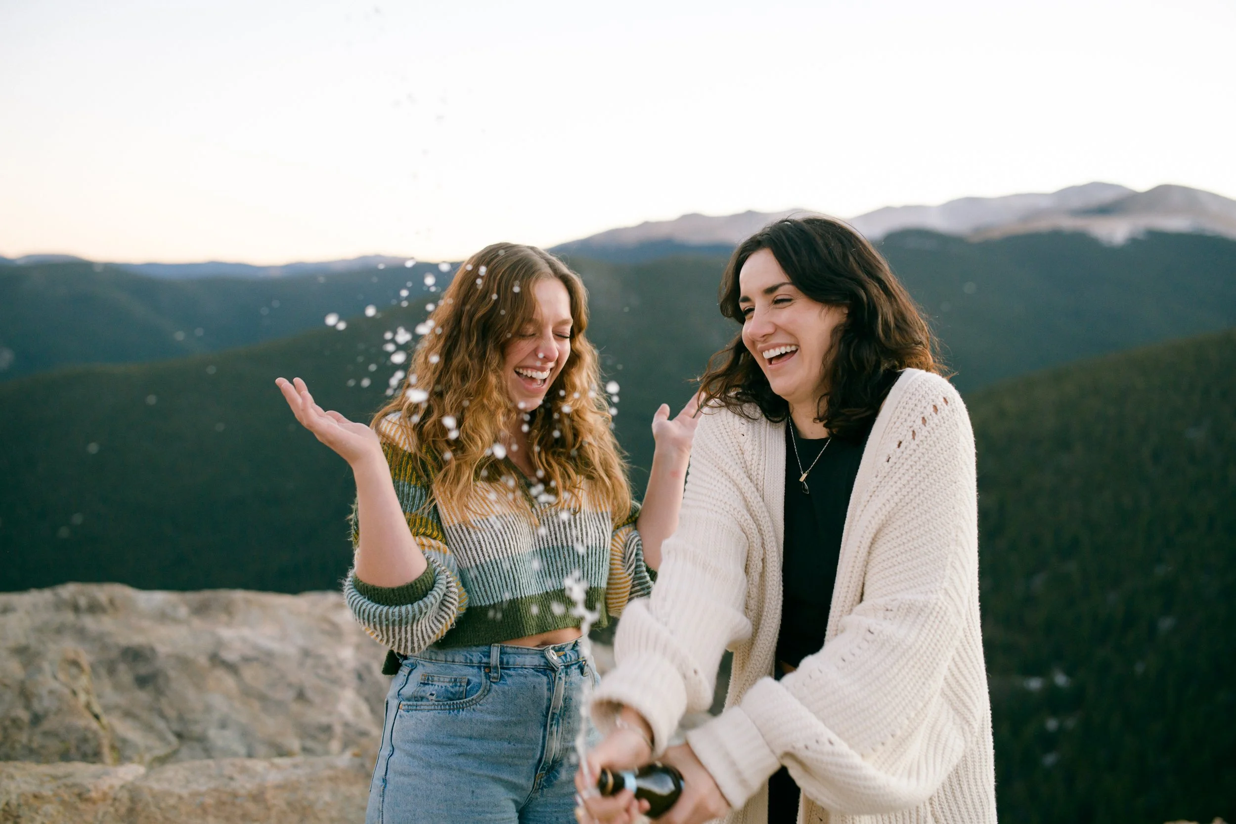 Two women celebrate their LGBTQIA+ queer colorado marriage proposal outdoors. One opening a bottle and the other spraying champagne with Juniper Pass, a mountain pass near Idaho Springs, CO, in the background