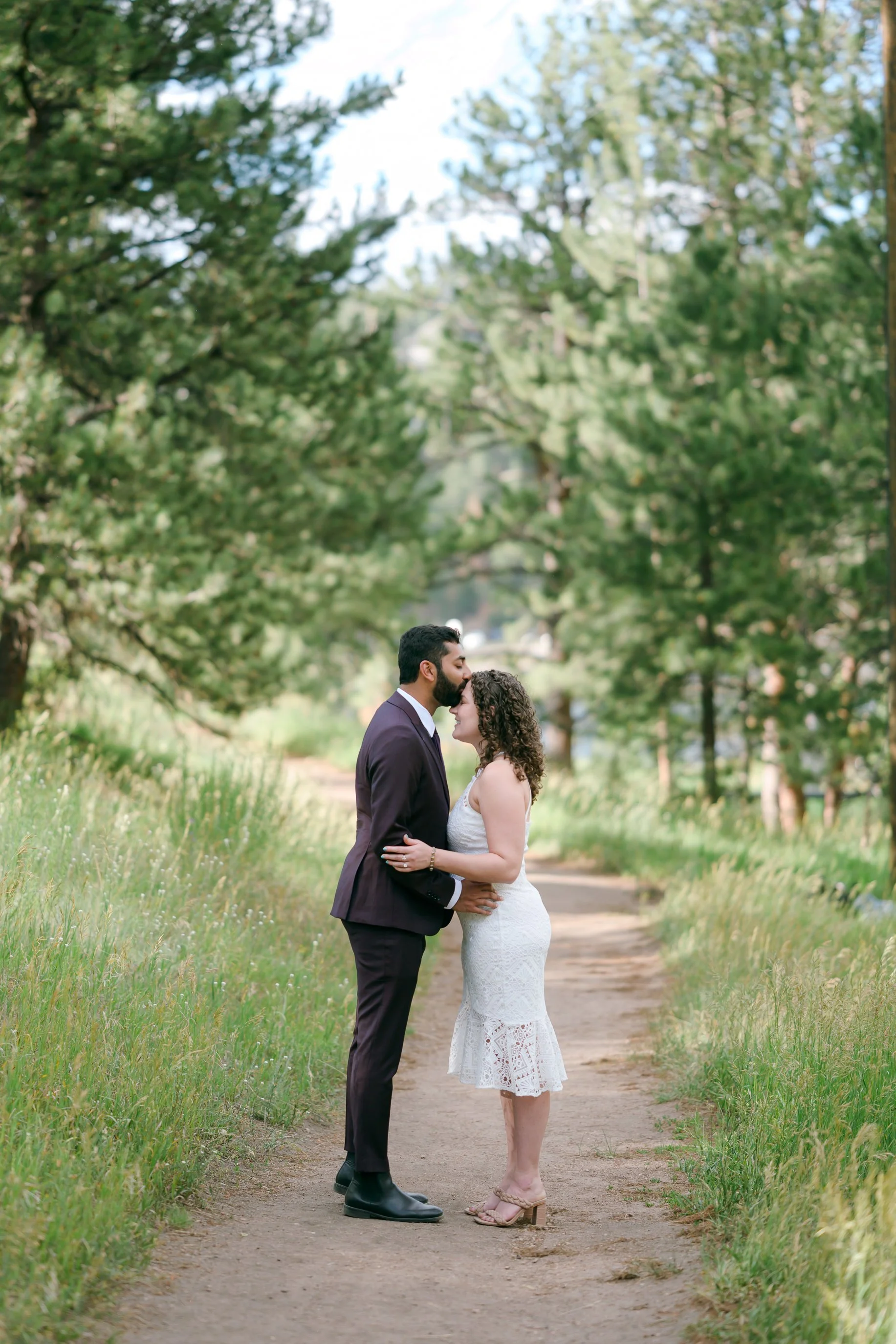 A BIPOC couple dressed in formal attire sharing a kiss on a dirt path surrounded by trees for their Evergreen Lakehouse engagement photos