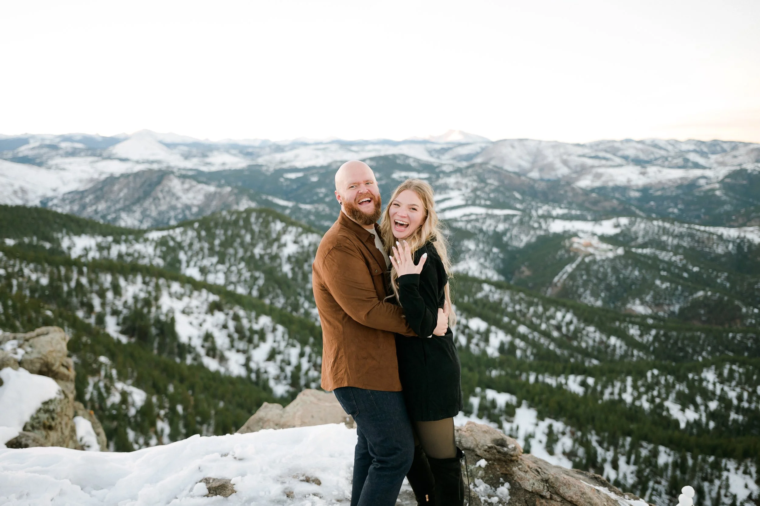 A happy couple embracing on a snowy mountain with snow-covered peaks and evergreen trees in the background for their boulder proposal photos