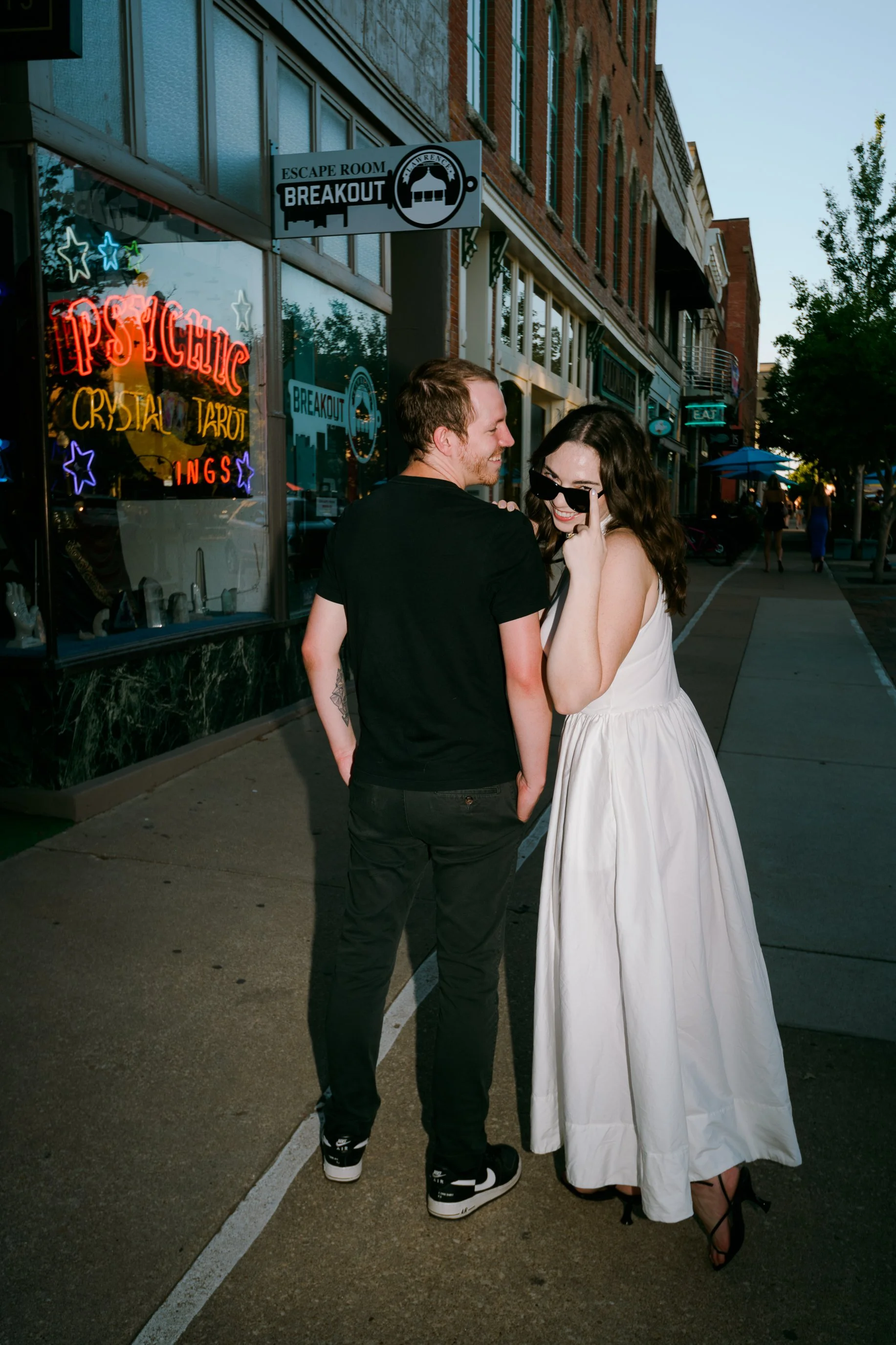 An engaged couple cuddles for a documentary style photography moment with flash photography while walking on a sidewalk on Mass Street a building with neon signs, smiling and chatting at dusk in Lawrence, Kansas.