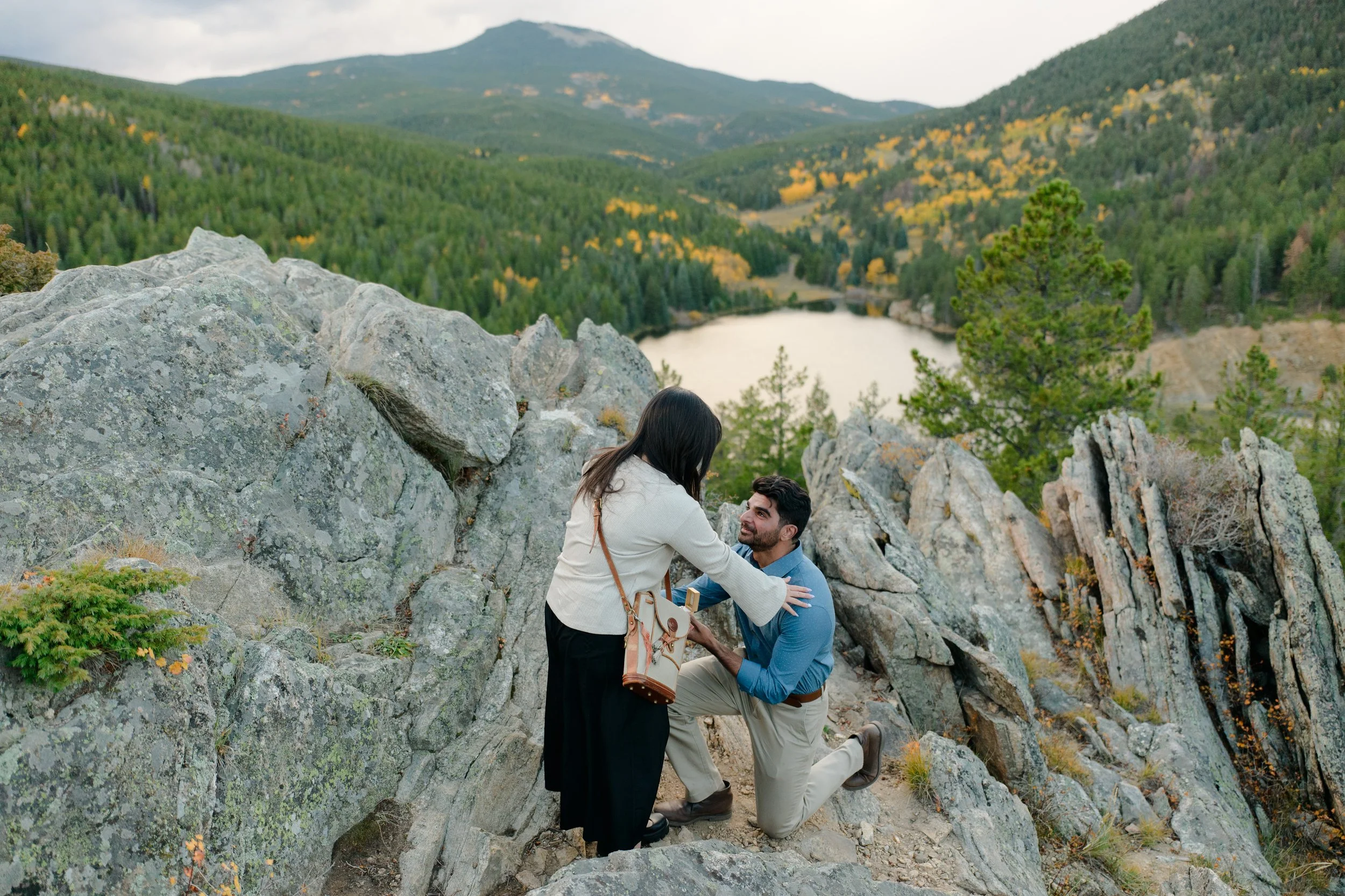 Indian man proposes to Asian woman with yellow aspens and a watershed in the background in Evergreen Colorado on a rock face