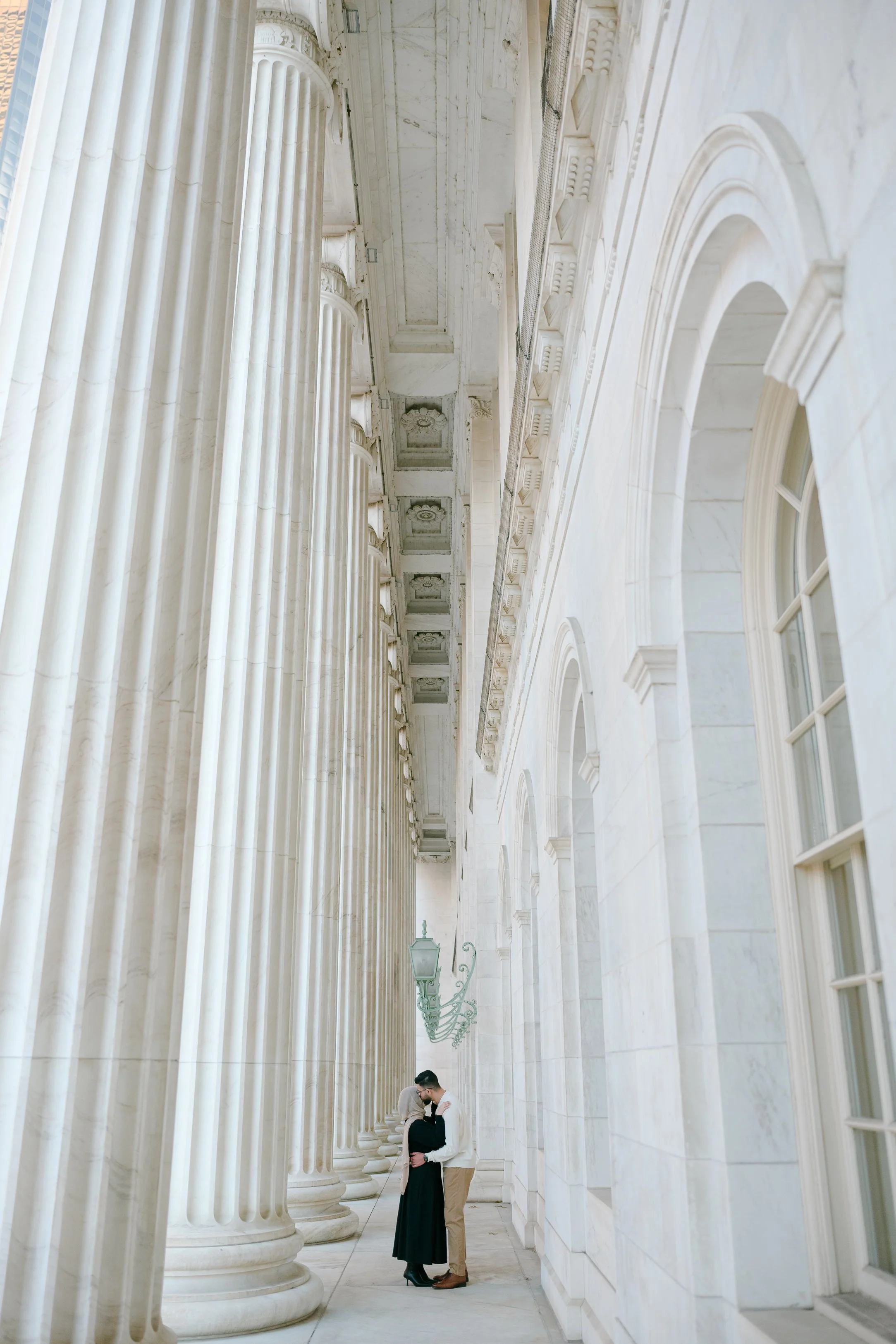 A BIPOC LGBTQIA+ couple stands close together in front of tall, white marble columns for their Byron White Court House Denver elopement photos. The couple is very small compared to the pillars