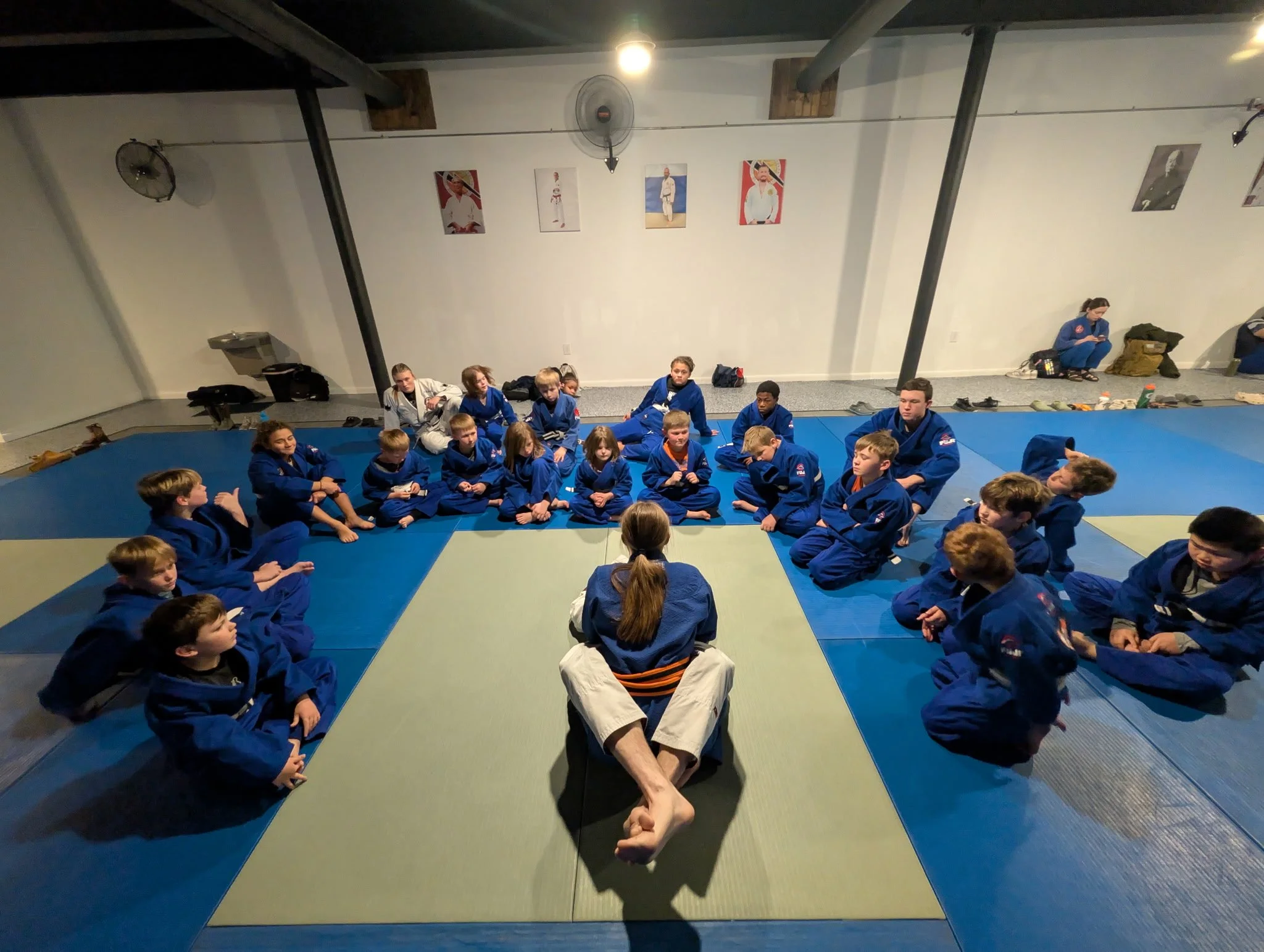 A group of young martial arts students sitting cross-legged on blue and green mats in a dojo, dressed in blue gis, with adults seated behind them. The dojo has white walls decorated with posters of martial artists, fans on the ceiling, and personal belongings on the floor.