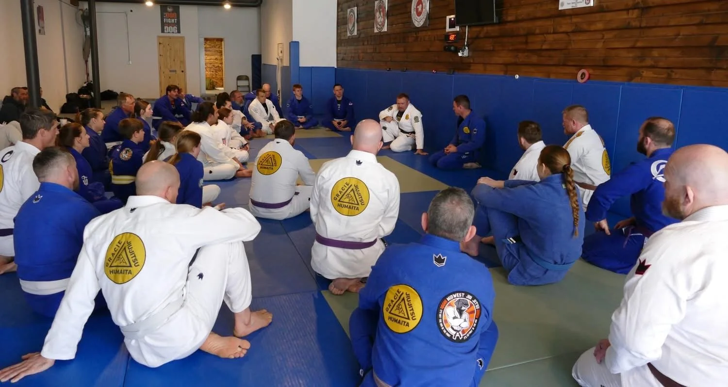 Group of people in Brazilian Jiu-Jitsu gis seated in a circle on a mat, listening to an instructor during a class or seminar in a martial arts gym.