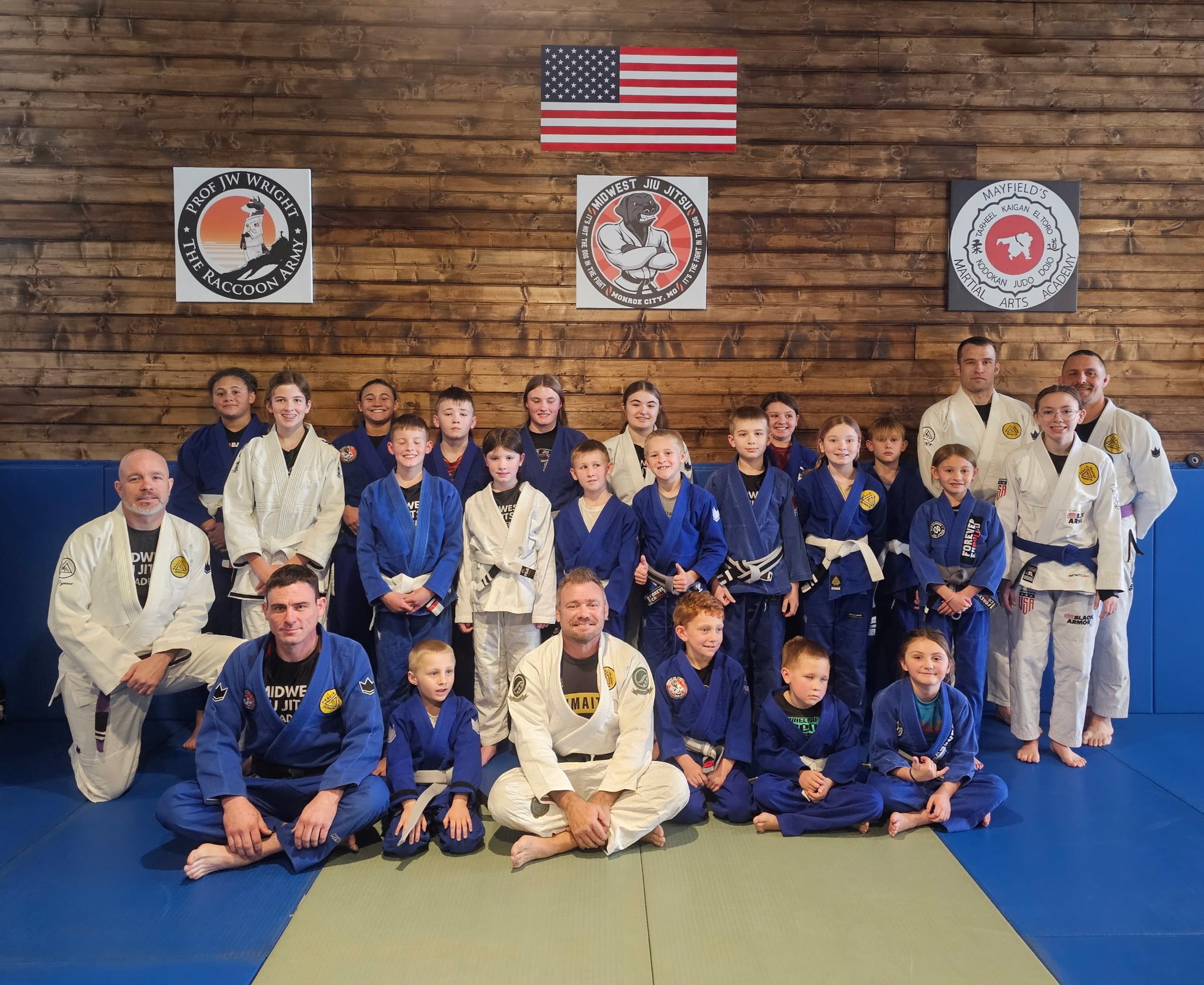 Group photo of children and instructors in martial arts uniforms at a dojo with American flags and martial arts banners on the wooden wall.