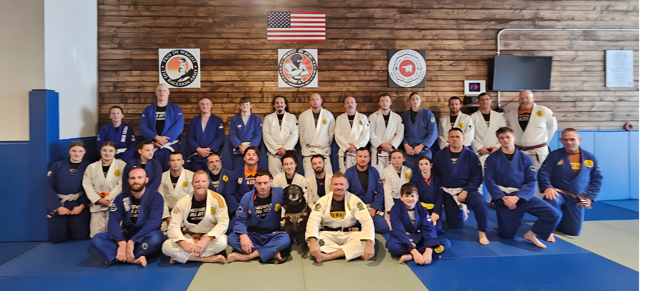 Group of Jiu-Jitsu practitioners, including children and adults, posing together in Martial Arts uniforms on a training mat in a dojo. The background features a wooden wall with martial arts logos and an American flag.