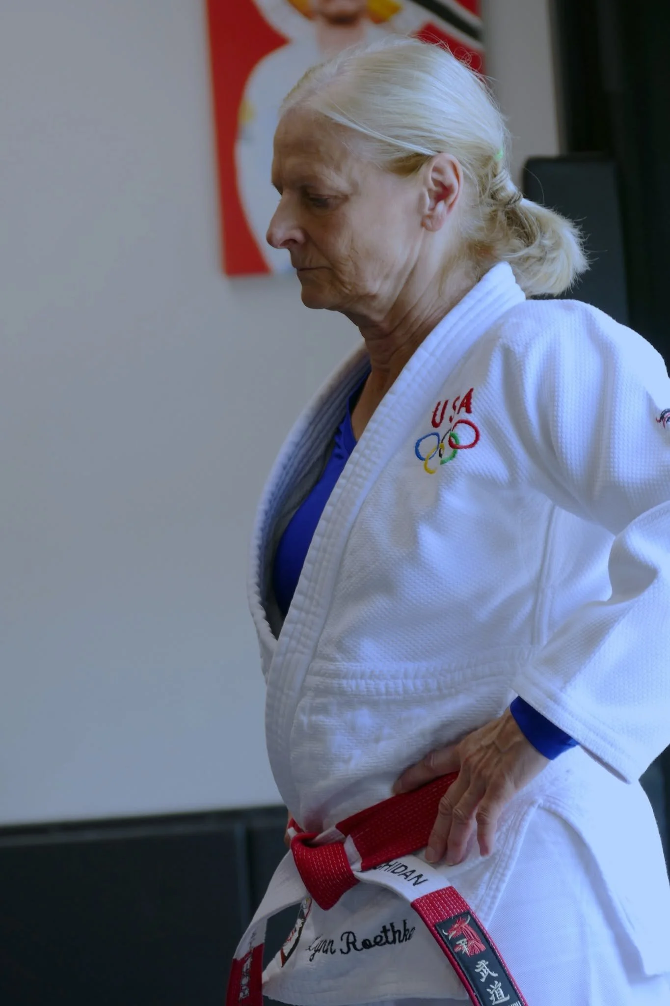 An elderly woman in a martial arts uniform standing with her hand on her hip. She has long gray hair tied back and is wearing a white gi with red and black belt. The background features a poster of a martial artist and the USA Olympic logo.