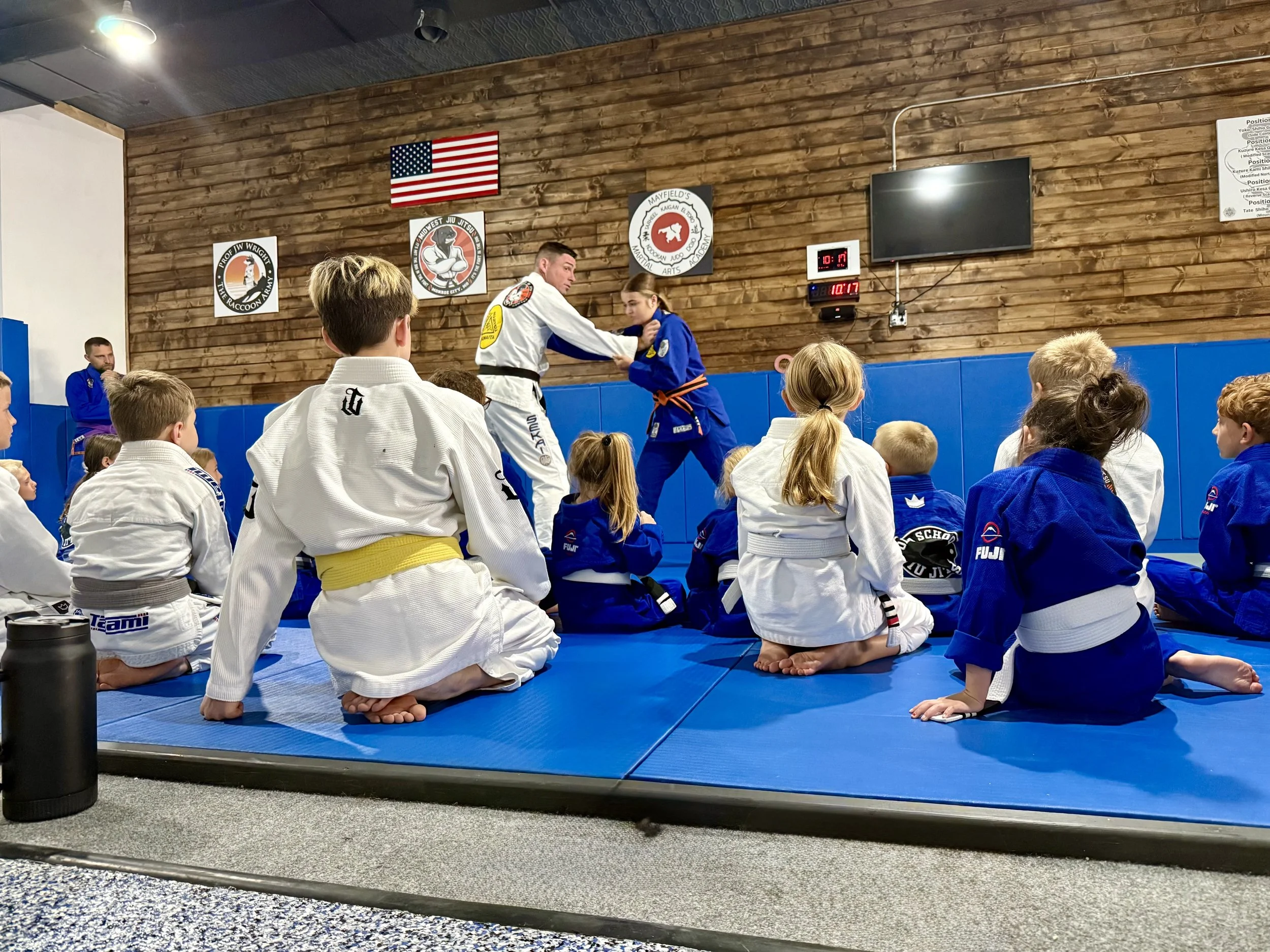 Martial arts class with children dressed in gi, sitting on blue mats, listening to instructors demonstrating techniques. Wooden walls, American flag, and martial arts banners are visible. Digital clock shows 10:17.