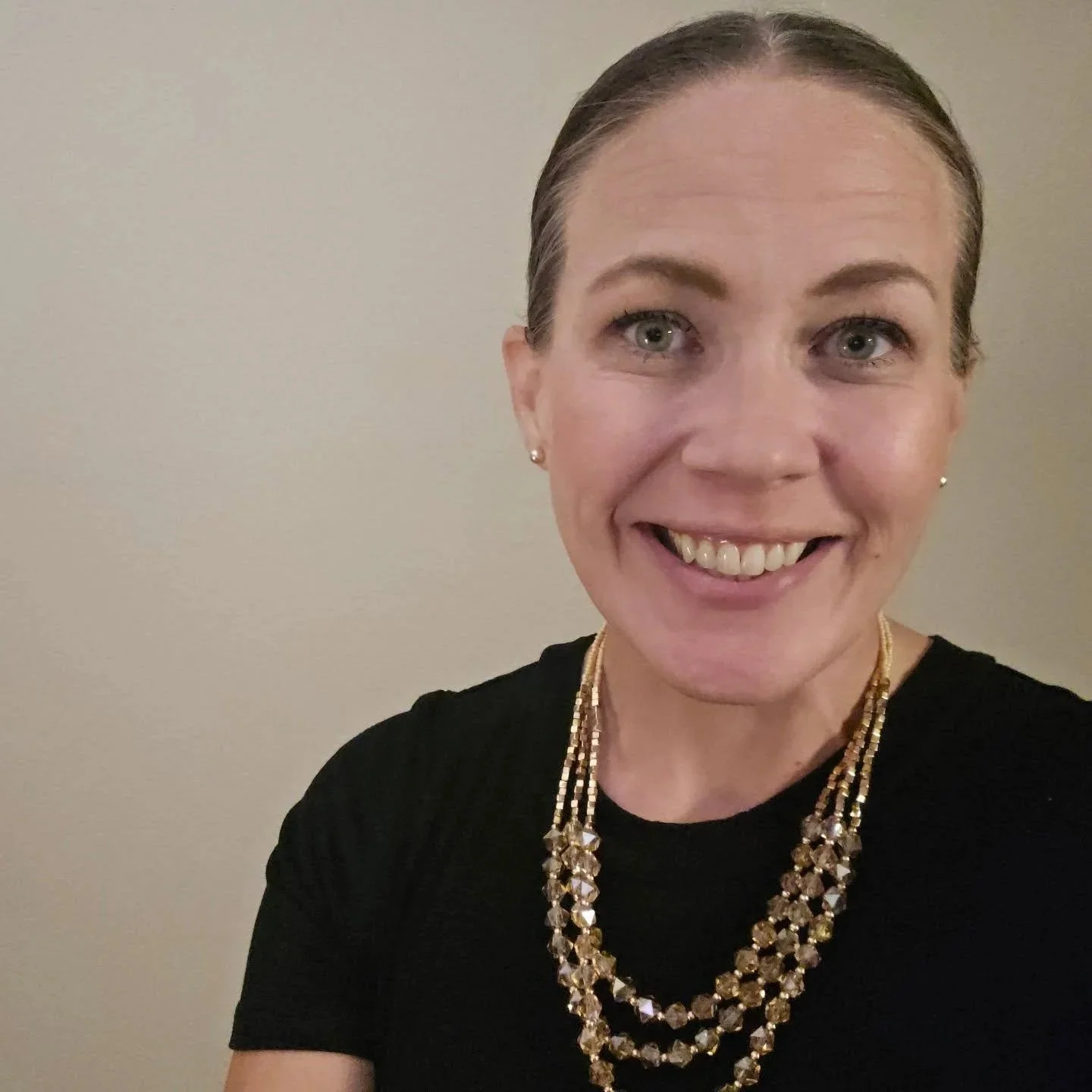 A woman smiling with blonde hair tied back, wearing pearl earrings, a black shirt, and a gold necklace, standing against a plain wall.