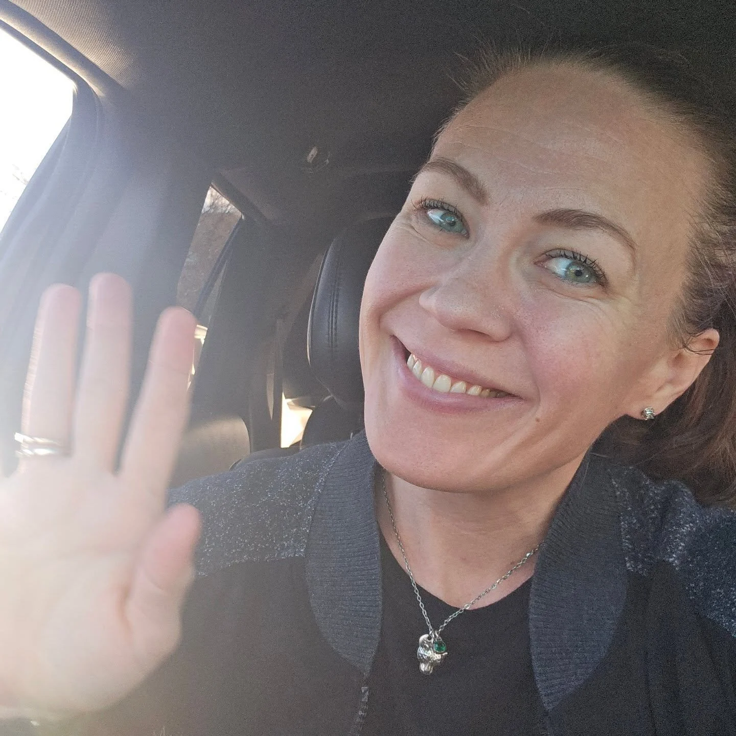 A woman with light skin, blue eyes, and brown hair smiling and waving at the camera while sitting in a vehicle.