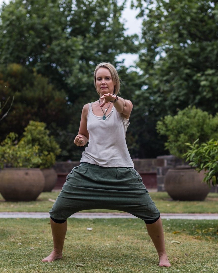 Woman practising Eight Sections of Brocade Qigong outdoors in a wide, grounded stance.