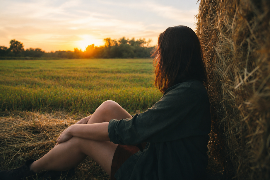 Person sitting beside a hay bale in a field at sunset, looking across the landscape