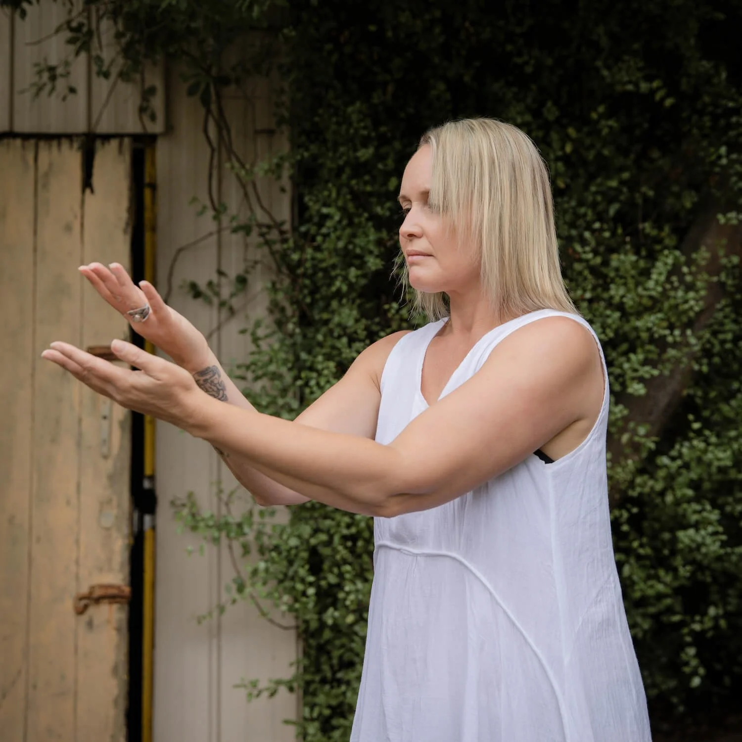A woman with blonde hair, wearing a white sleeveless dress, holding her hands out in front of her as if performing a gesture outdoors next to a wooden fence and green foliage.