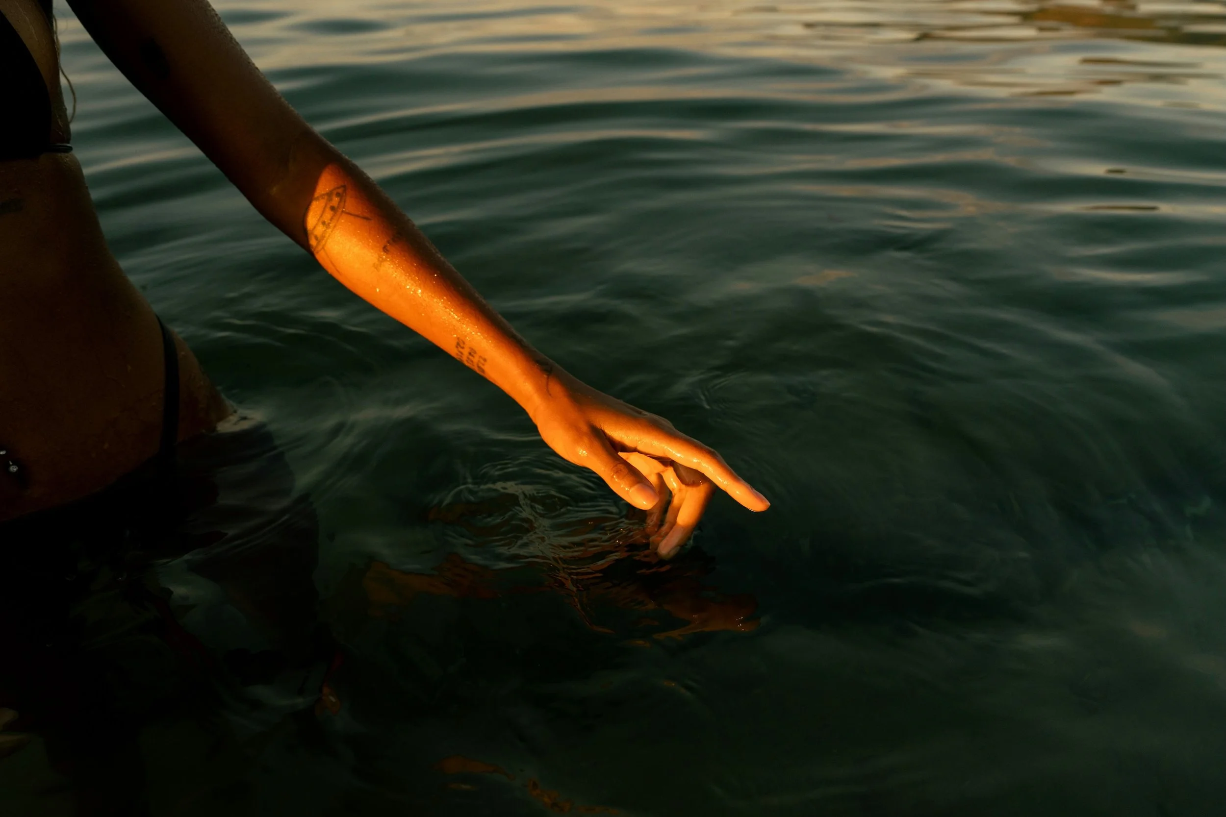 Person reaching one hand into still water, creating gentle ripples across the surface in warm evening light.