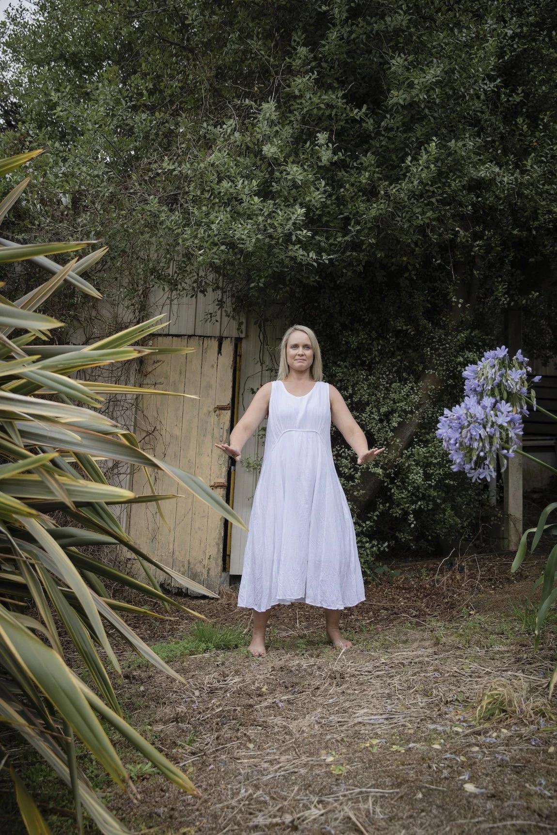 Woman practising Qigong barefoot outdoors in a garden, standing among plants with her arms gently open at her sides.