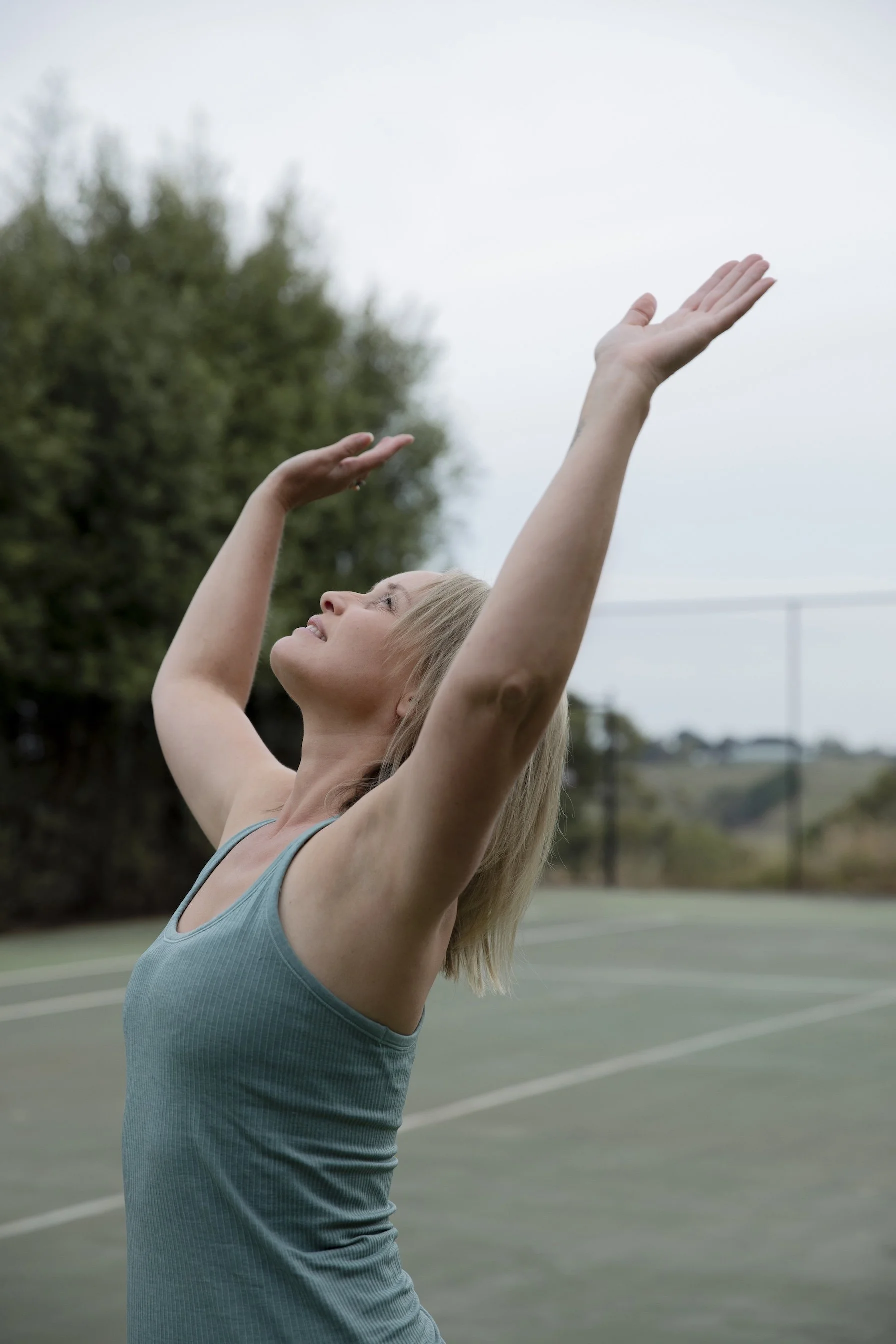 Woman practising a flowing Qigong movement outdoors, with both arms raised and an open sky behind her.