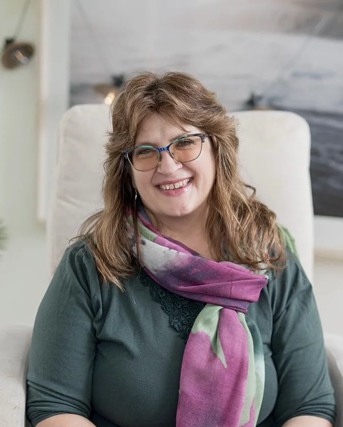 A smiling woman with wavy brown hair and glasses, sitting on a beige armchair, wearing a floral blouse and purple cardigan.