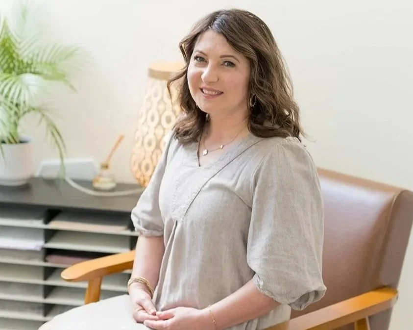 A woman with brown hair and light skin smiling, sitting on a beige couch in front of a large green plant and a wooden wall, wearing a dark blue blouse, a gold watch, and a gold chain necklace.