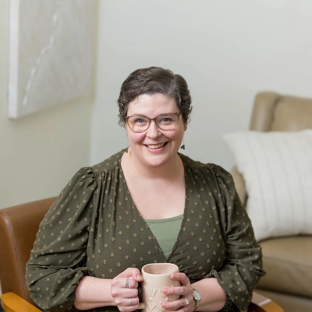 A woman with short dark hair, glasses, and hoop earrings sits on a beige armchair, smiling at the camera. She wears a patterned blouse with ruffles and a black top underneath, with a ring on her finger. In the background, there is a side table with decorative items and a lamp, and sheer curtains covering a window.